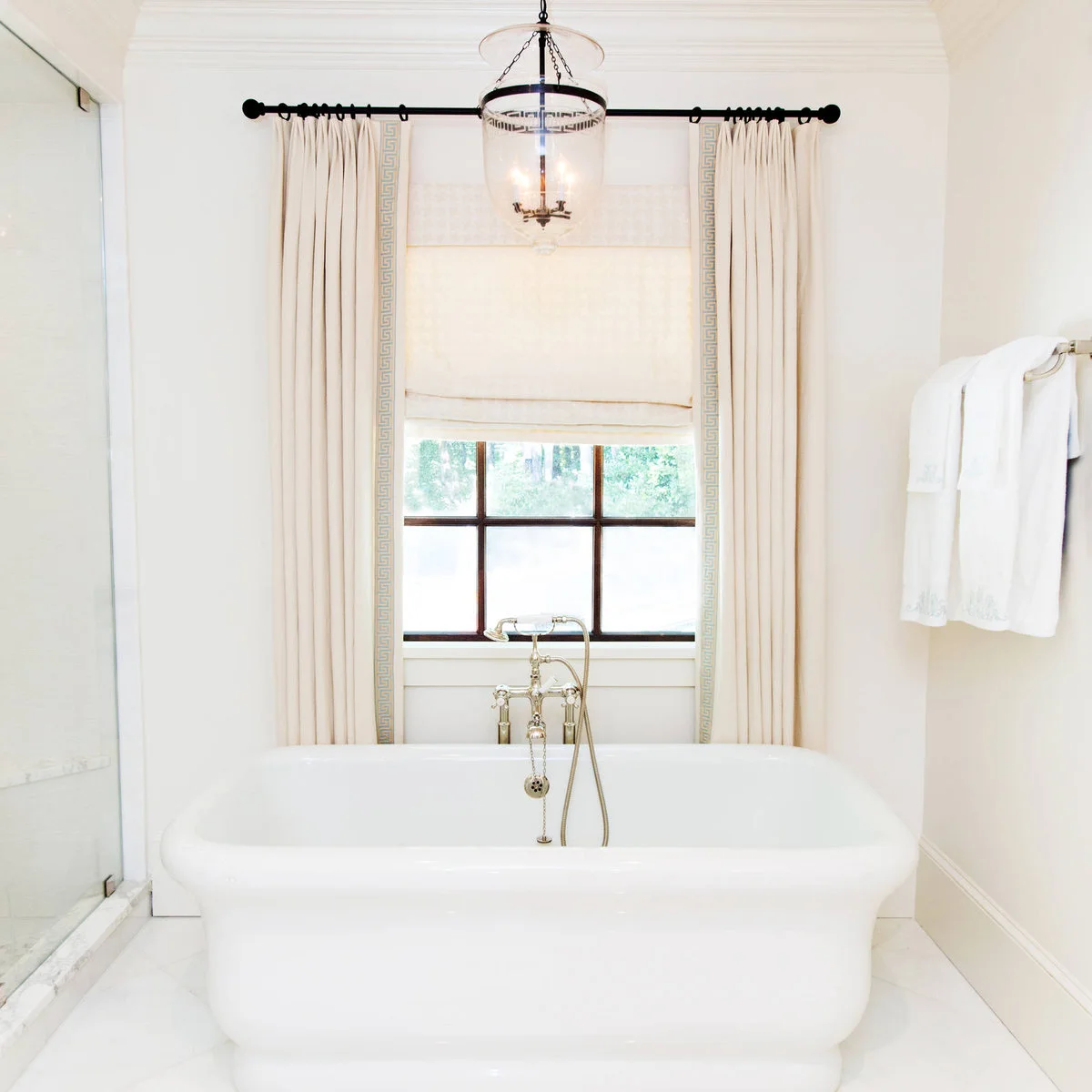 Cream curtains with a pale blue geometric border frame a shaded window behind a white bath.