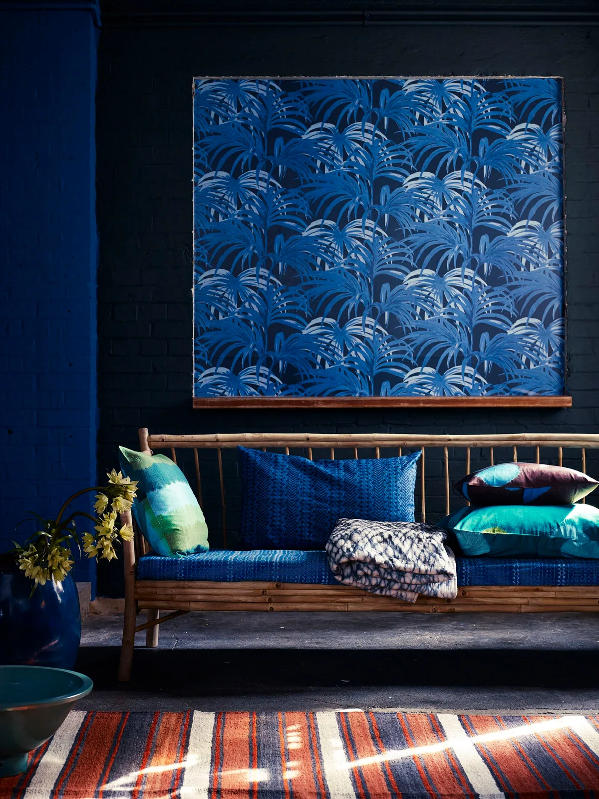 A rectangular blue leaf-patterned wallpaper panel hangs above a wood-framed sofa against a dark wall.