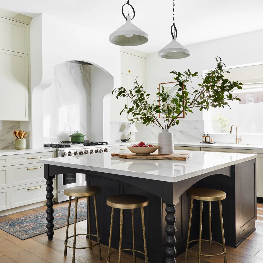 A dark island with turned legs and a pale counter faces an arched range alcove and light kitchen cabinets.