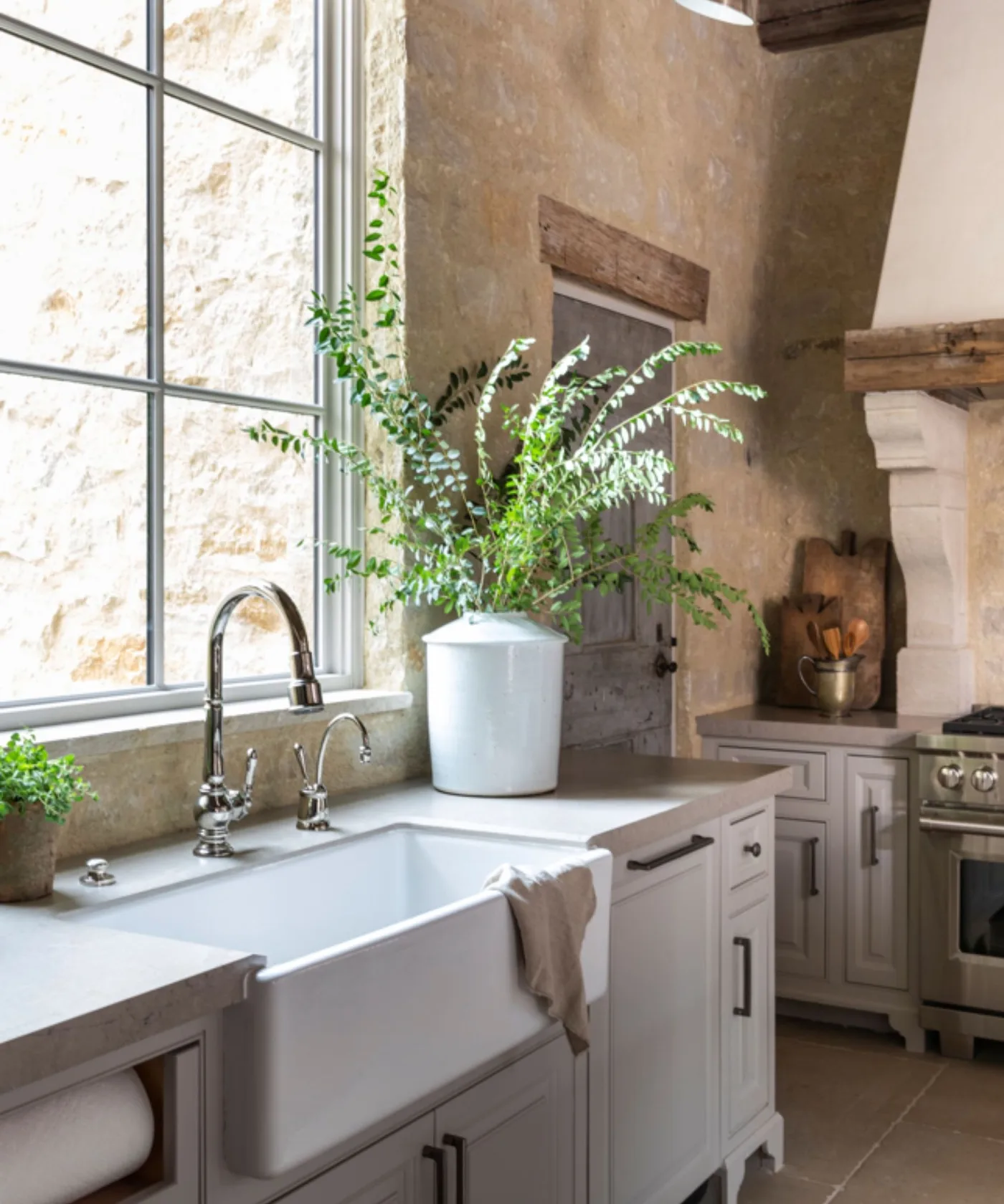 White apron-front sink beneath a gray-framed window, with gray cabinets, textured pale wall and jars beside a plant.