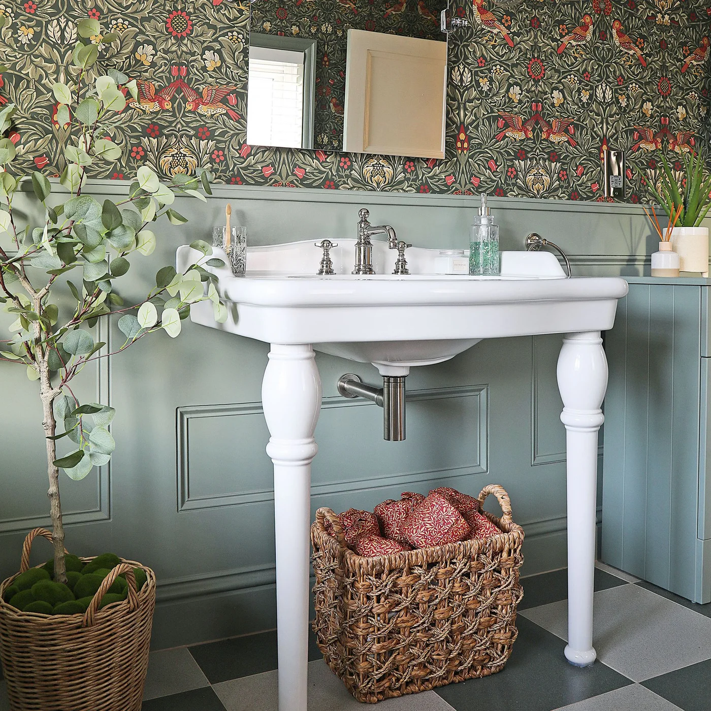 Sage paneling sits below richly patterned bird-and-leaf wallpaper behind a white console basin with a rectangular mirror and woven baskets.