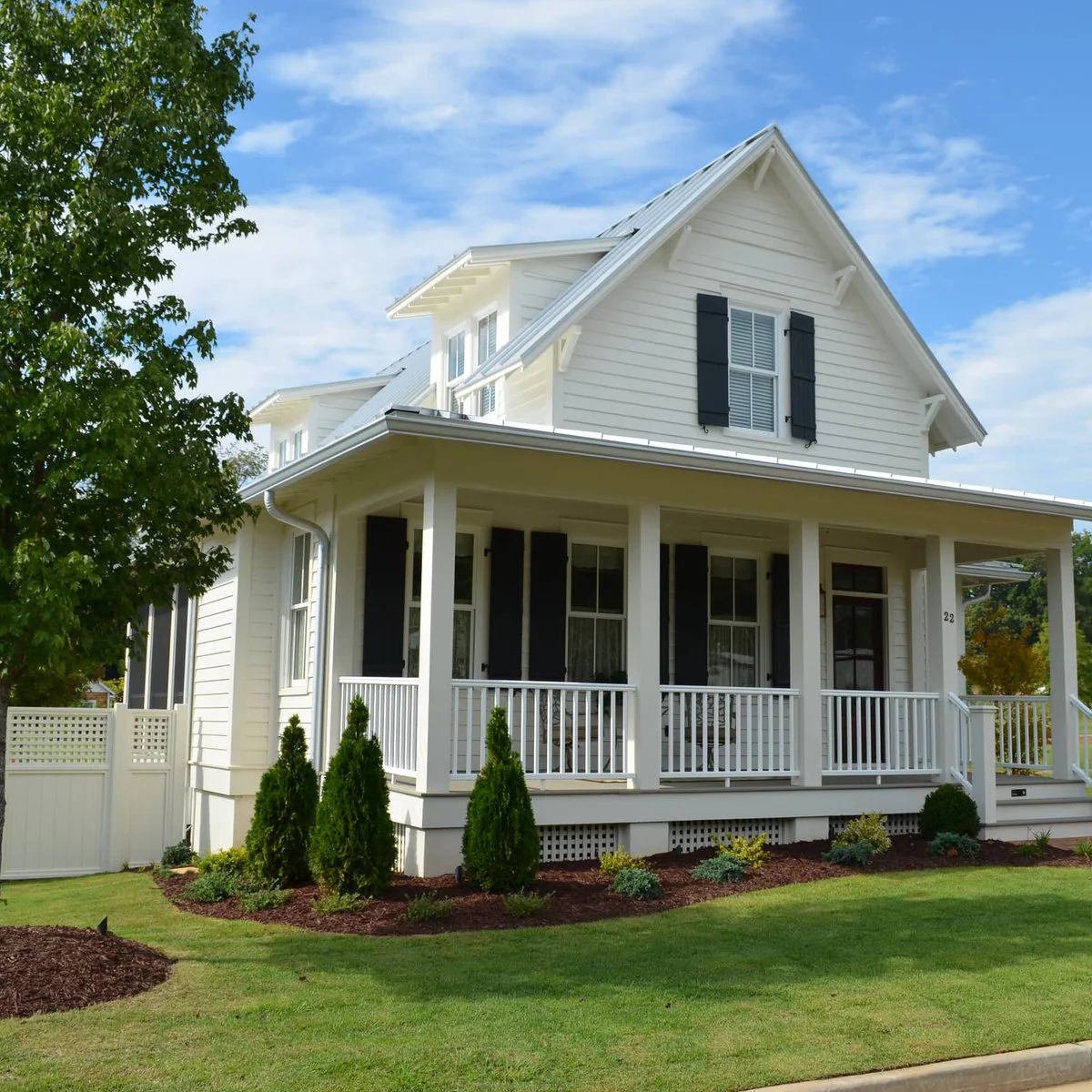A white wraparound porch with evenly spaced conical evergreens in a mulched bed below the railing.