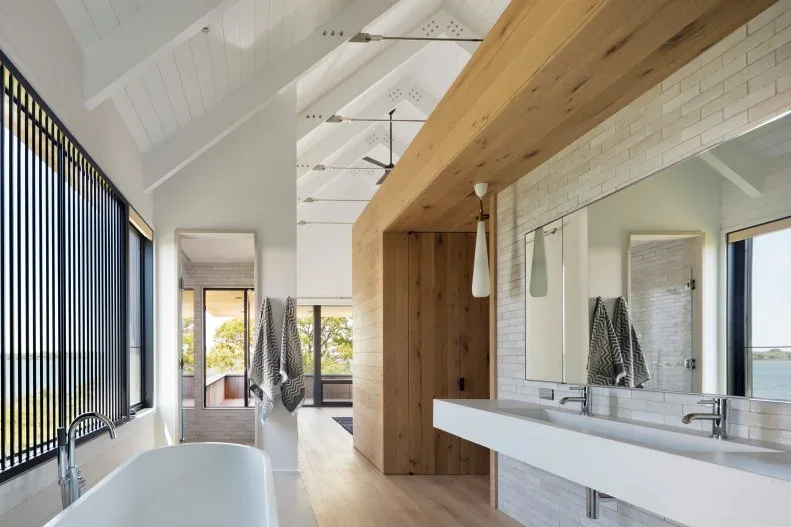 A long pale double washstand sits below a broad mirror beside a warm wood-lined wall opening, white ceiling beams and a tub near dark window lines