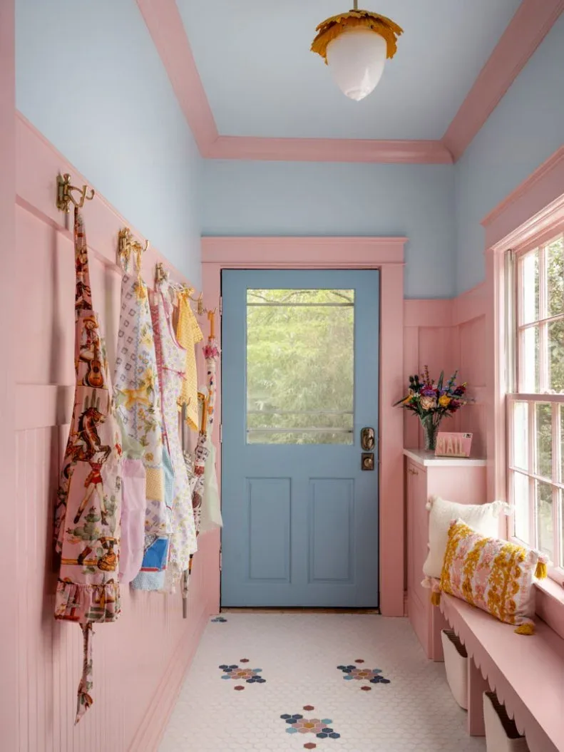 Entry mudroom with pink paneling and bench, blue glazed door, patterned hanging garments and yellow fringed cushion.