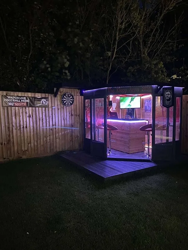 Small corner garden pub with an angled glazed entrance, two stools and purple evening lighting