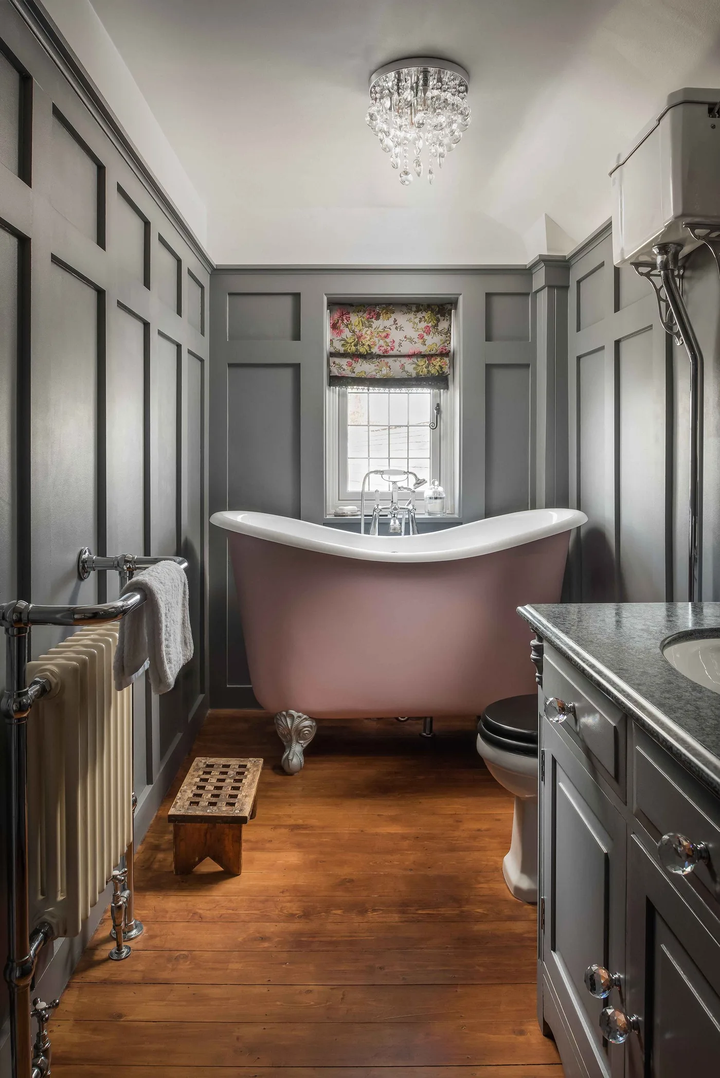 Narrow bathroom with a pink freestanding bath, gray paneled walls, floral window blind, dark vanity and wooden floor.