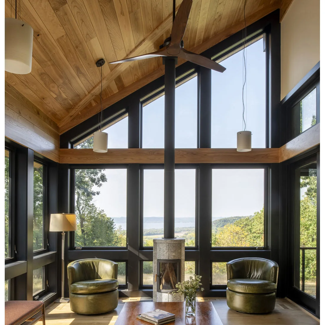 A small pale cylindrical stove and dark flue sit between green rounded chairs in a glazed room with a wood ceiling, dark frames and a low wood table.