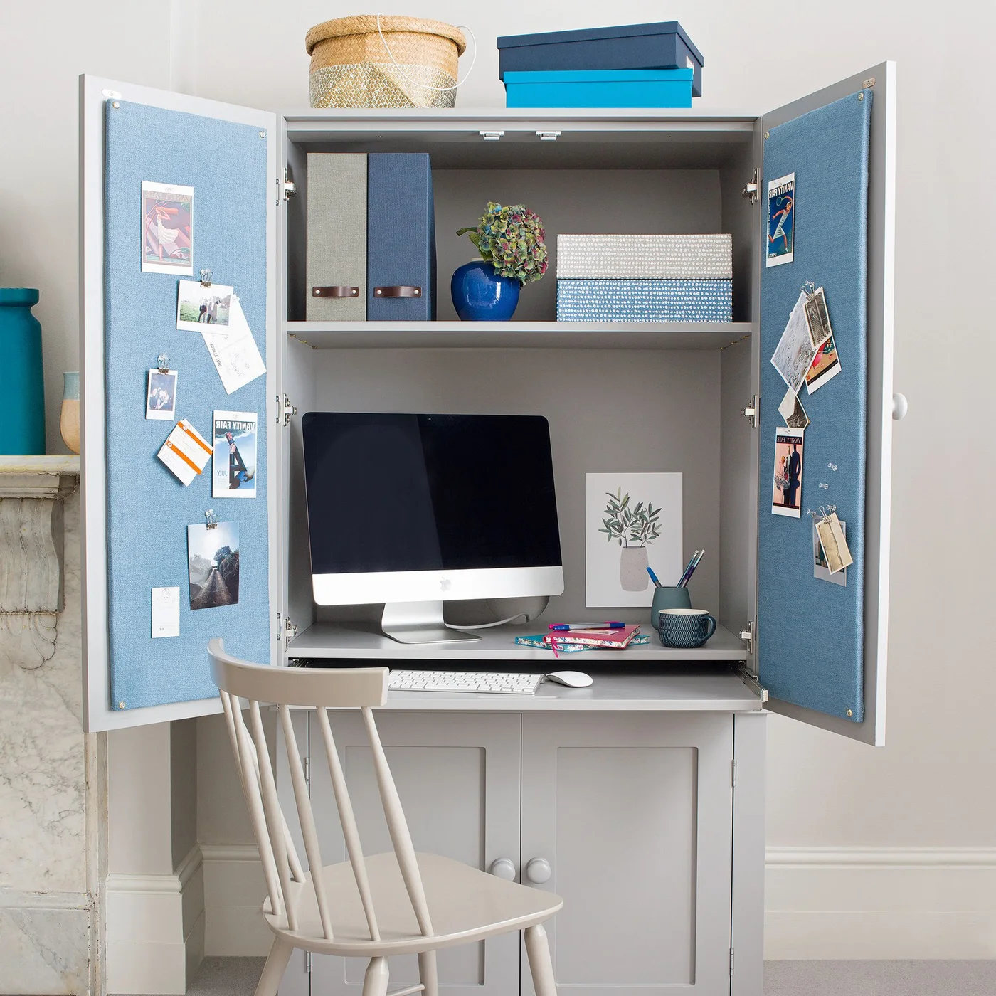 A gray work cabinet opens to reveal a monitor, keyboard shelf, storage boxes and blue pinboard panels on the inside of its doors.