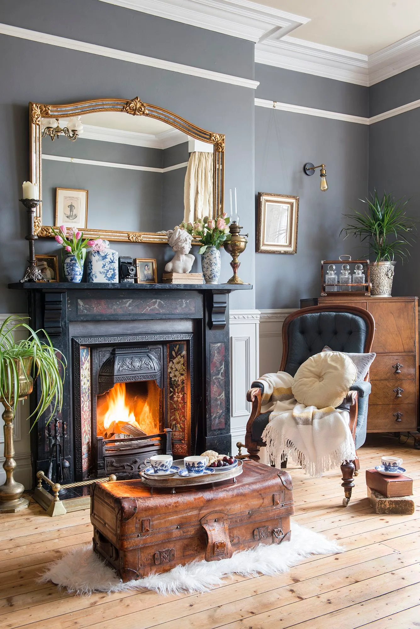 Gray room with dark ornate fireplace, large gold mirror, blue and white vessels, leather-look chair and trunk table