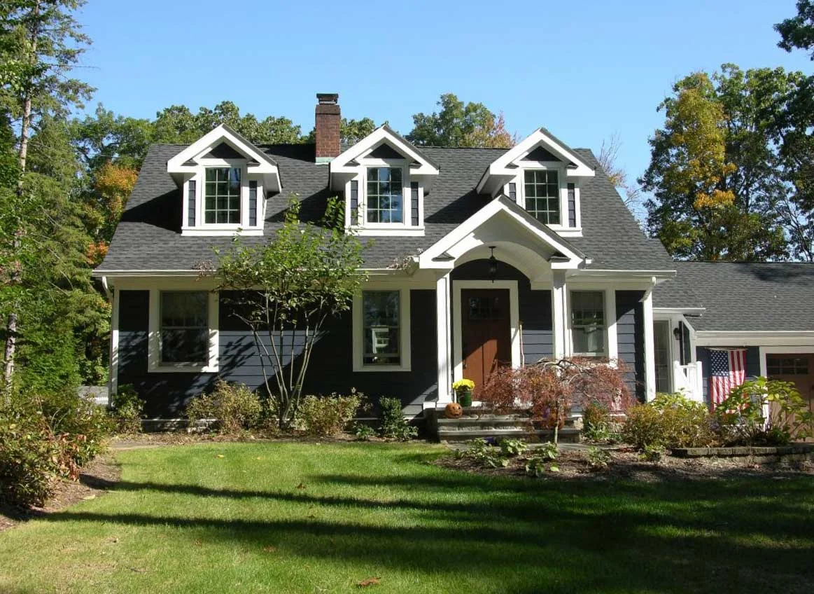 A dark blue Cape has three white-trimmed dormers, a central gabled porch entry and a lower roof extending toward the garage.