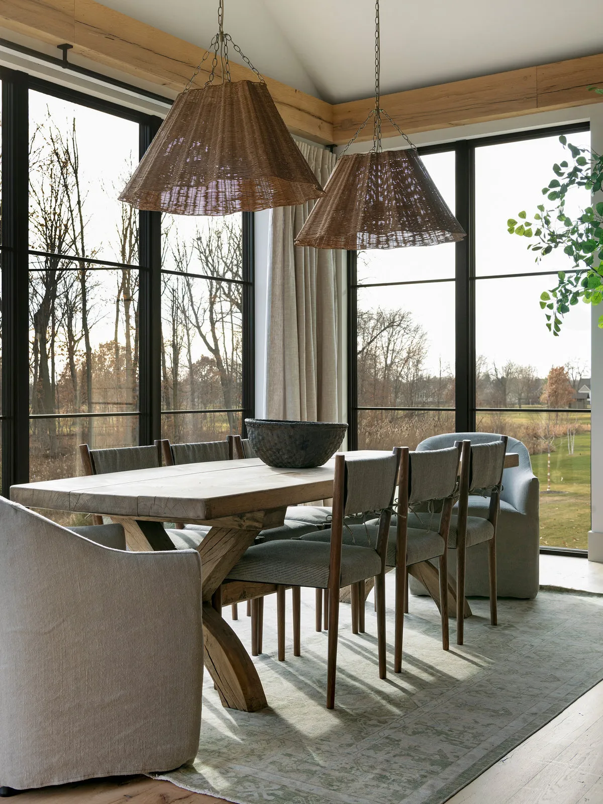 A pale trestle table stands beneath woven shades beside generous glazing.