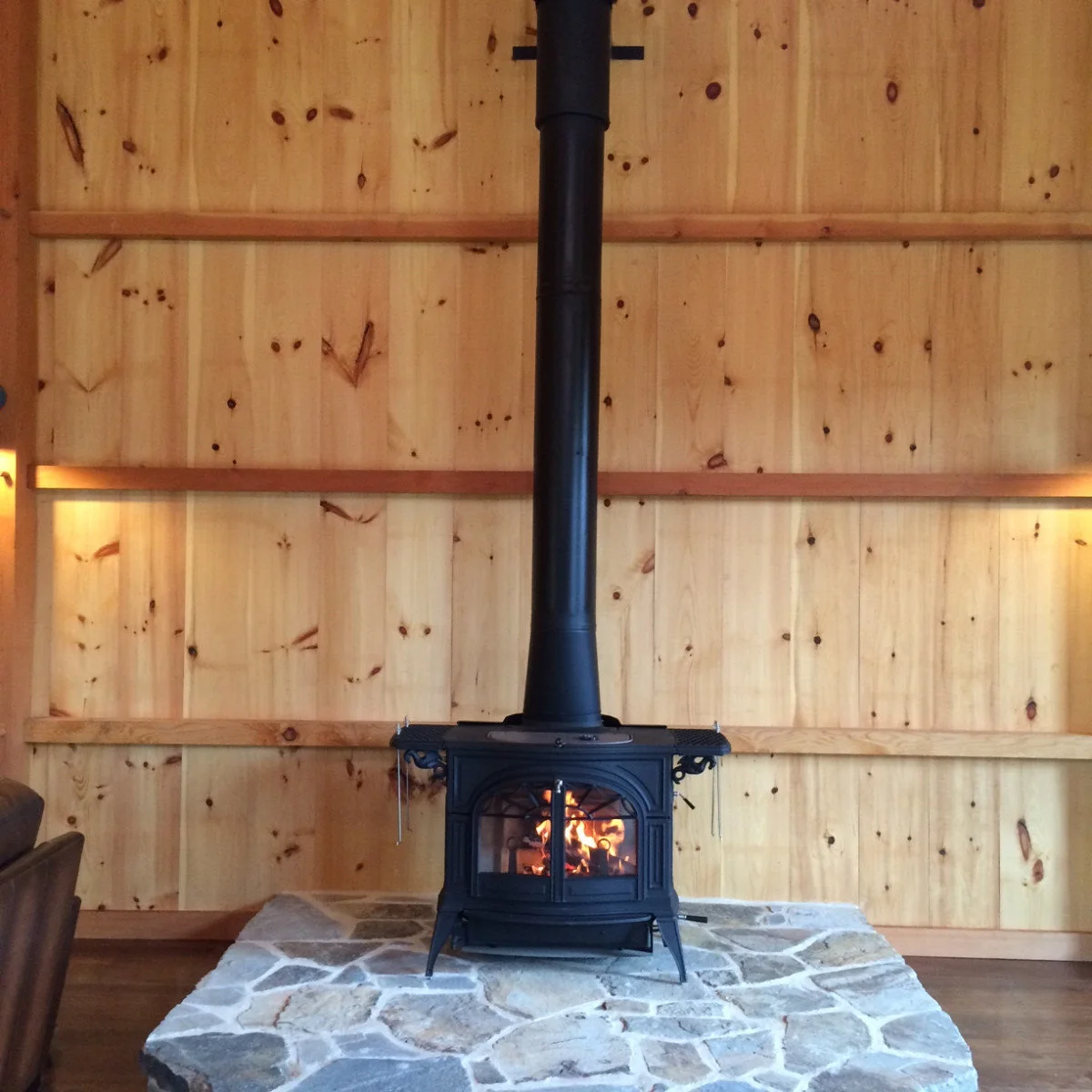 A black stove with an arched double-door front stands on a broad pale stone hearth against a plain timber-board wall.