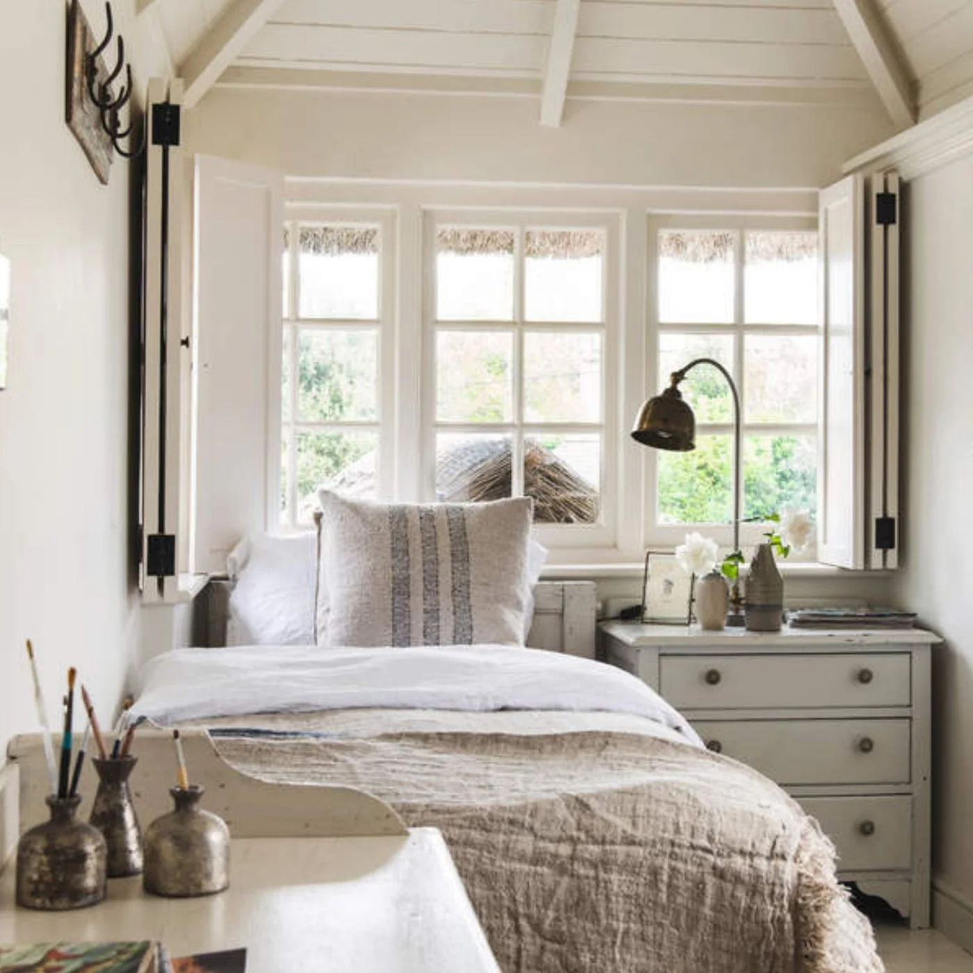 A single bed stands beneath three windows in a narrow pale bedroom beside a small drawer chest, with a long narrow desk on the opposite side.