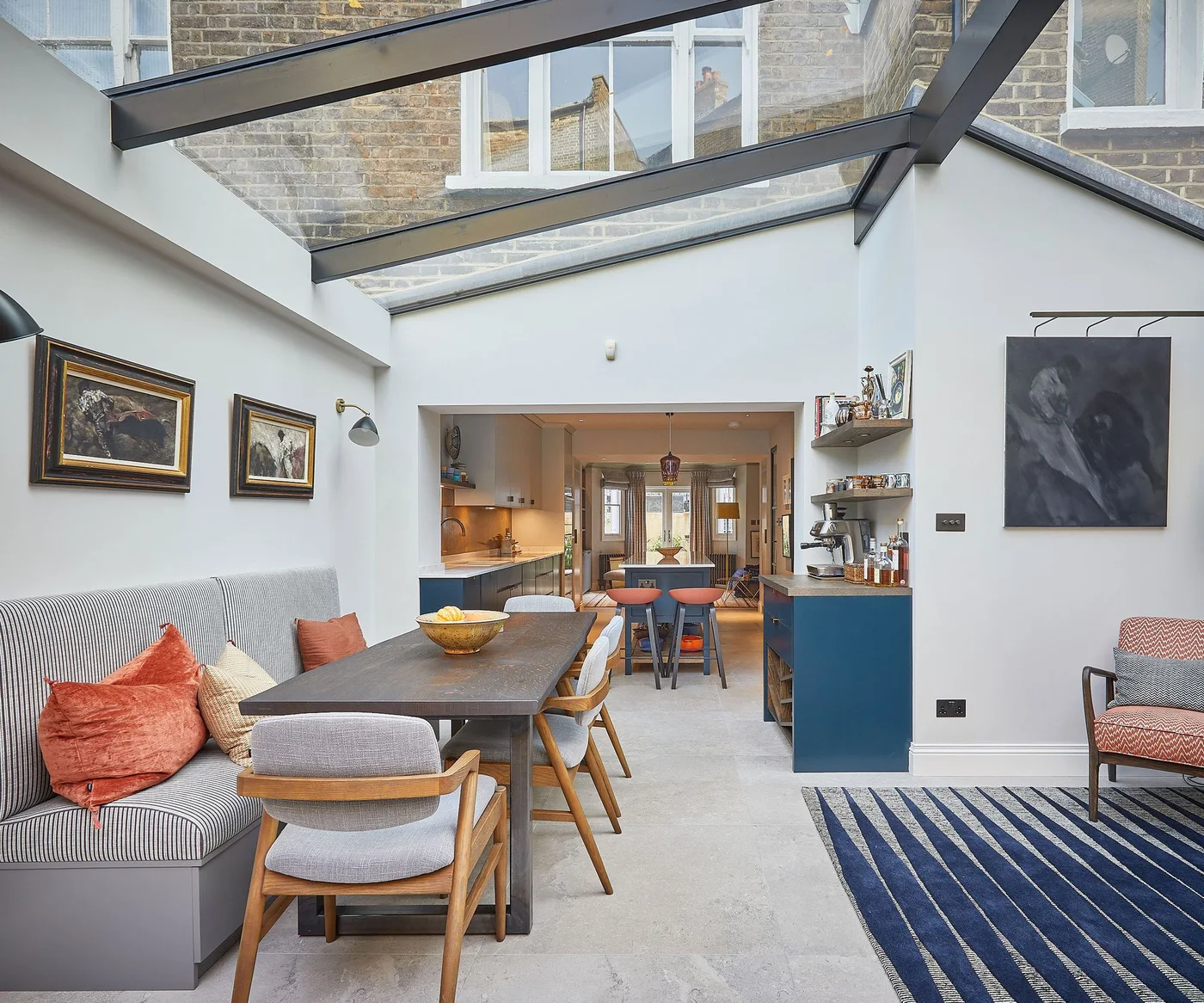 A long table, striped wall bench and wooden upholstered chairs occupy a glazed-roof area beside blue kitchen cabinetry