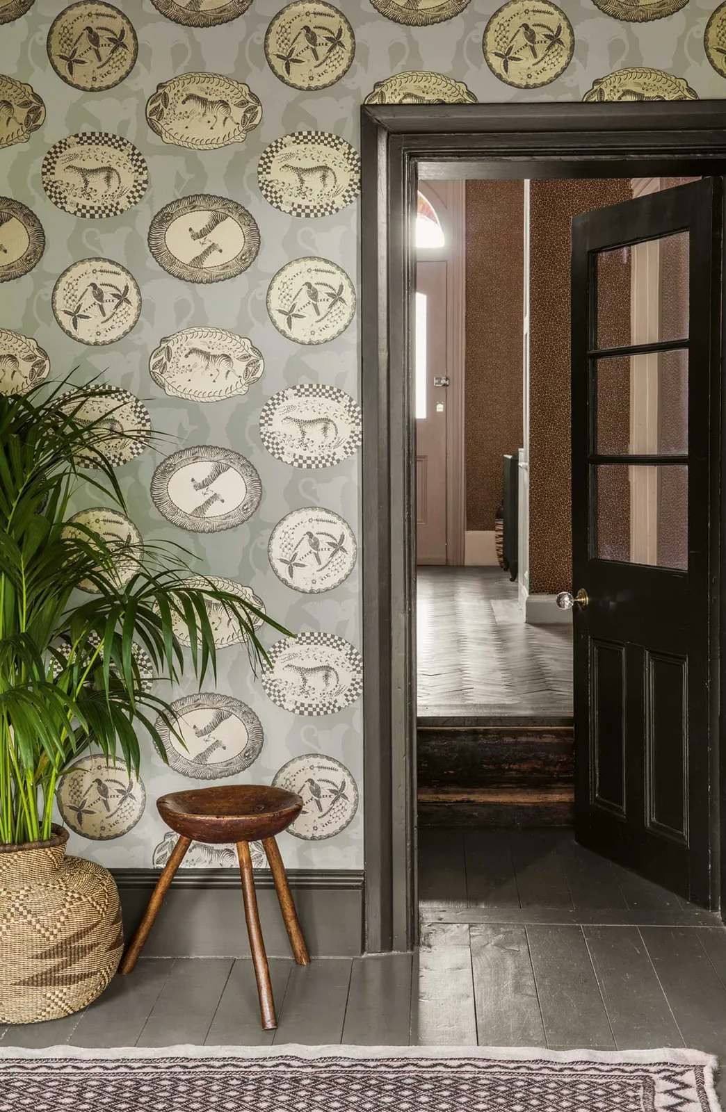 Illustrated round medallions repeat across a pale green hallway wall beside a dark glazed door, wooden stool and palm in a woven basket