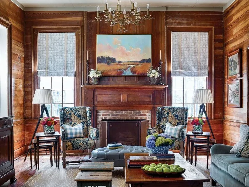 A timber mantel and brick inner surround sit among warm timber-paneled walls and patterned armchairs.