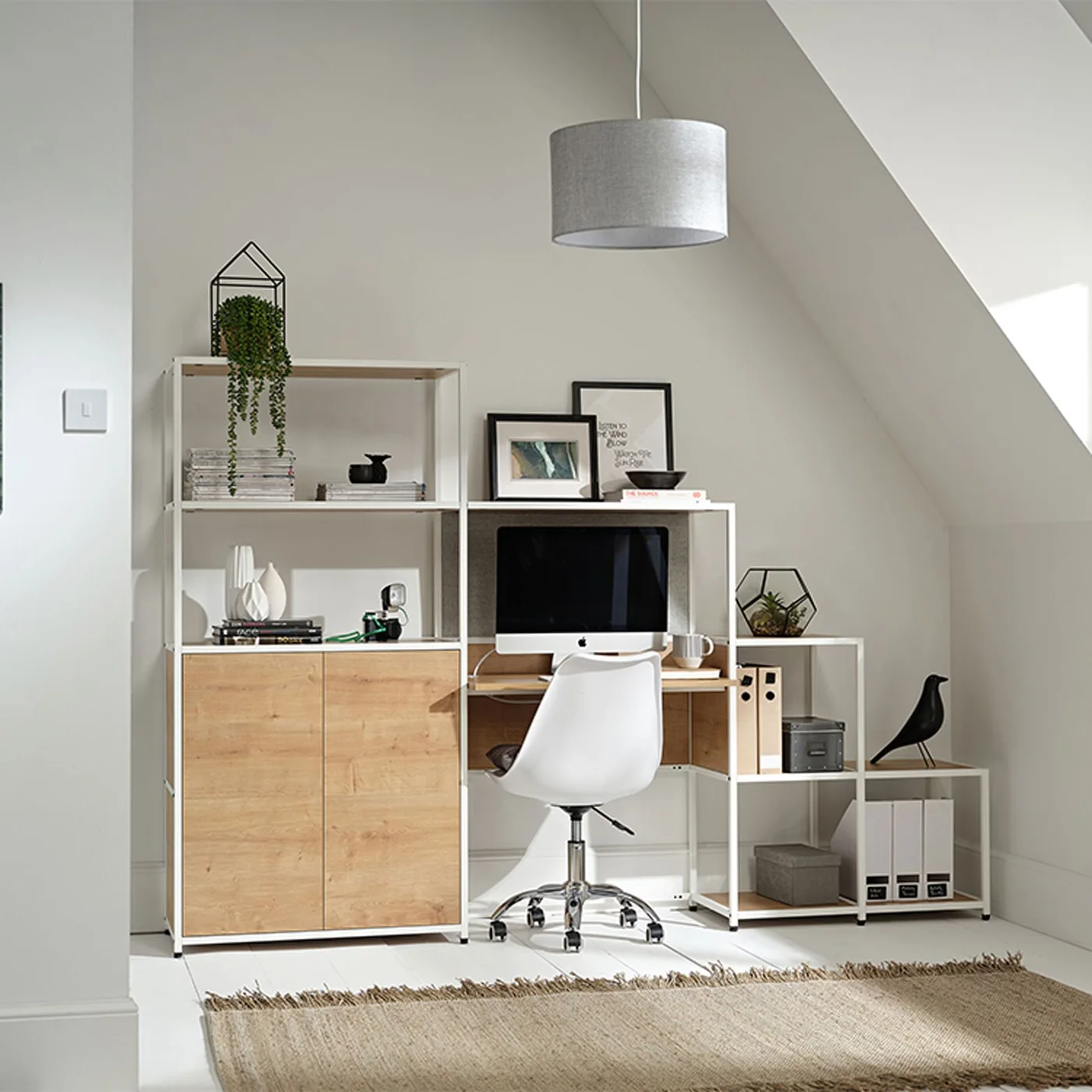 A white modular desk and shelving unit with wooden cupboard fronts sits against a pale gray wall beneath a gray pendant