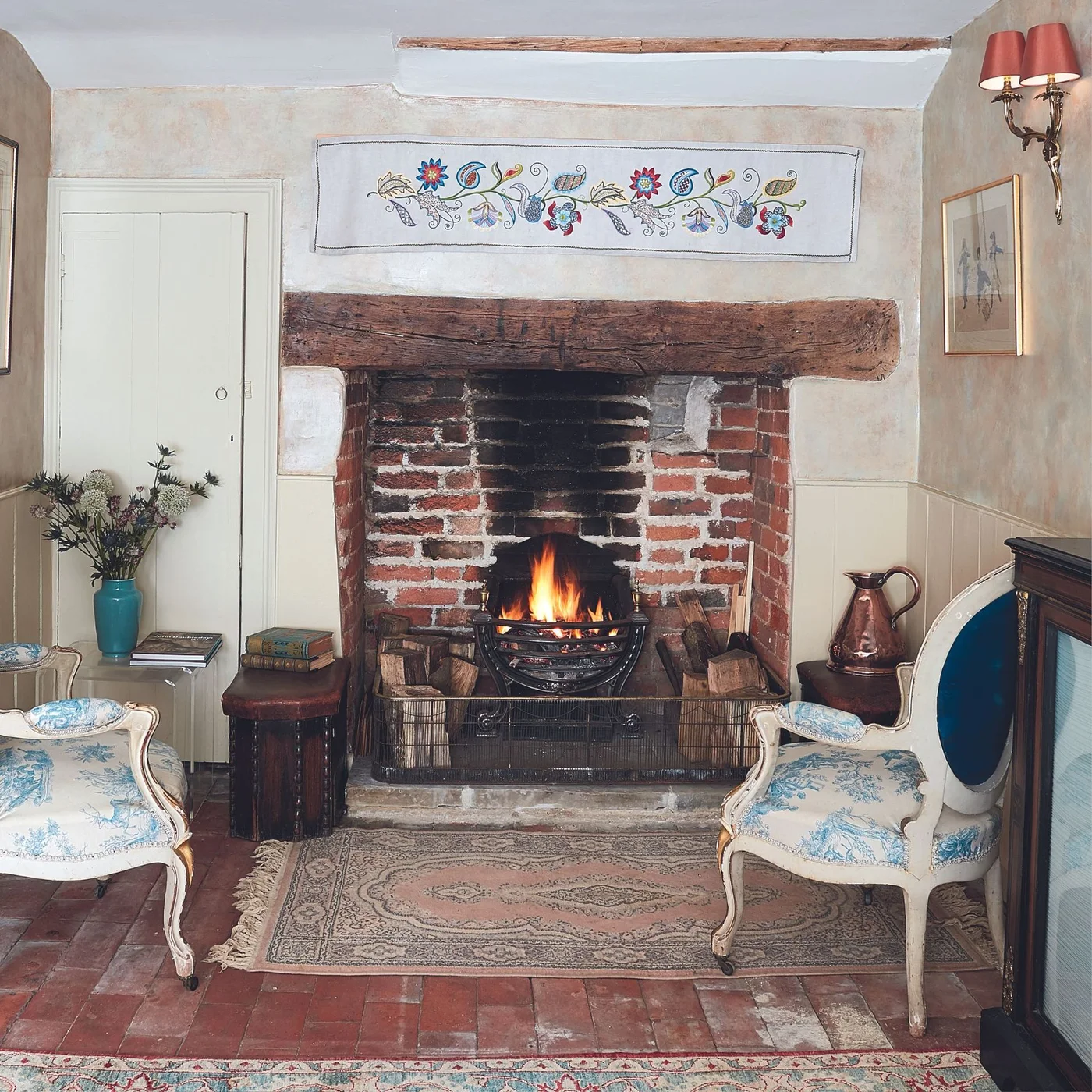A broad brick fireplace with a rough timber lintel sits beneath a decorative floral panel between pale walls and curved chairs.