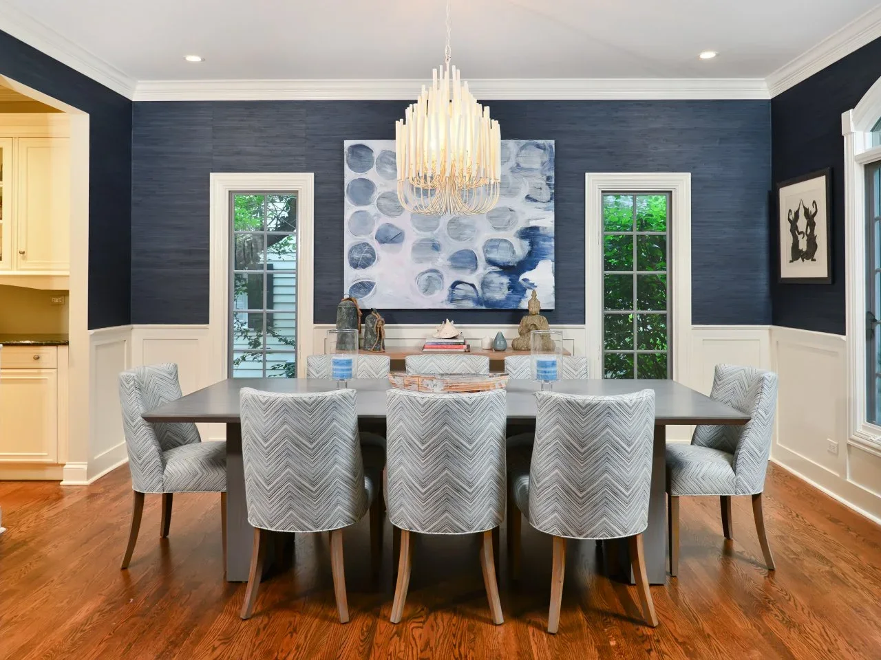 Textured navy upper walls and white lower paneling surround a gray dining table, pale patterned chairs and a long pale chandelier.