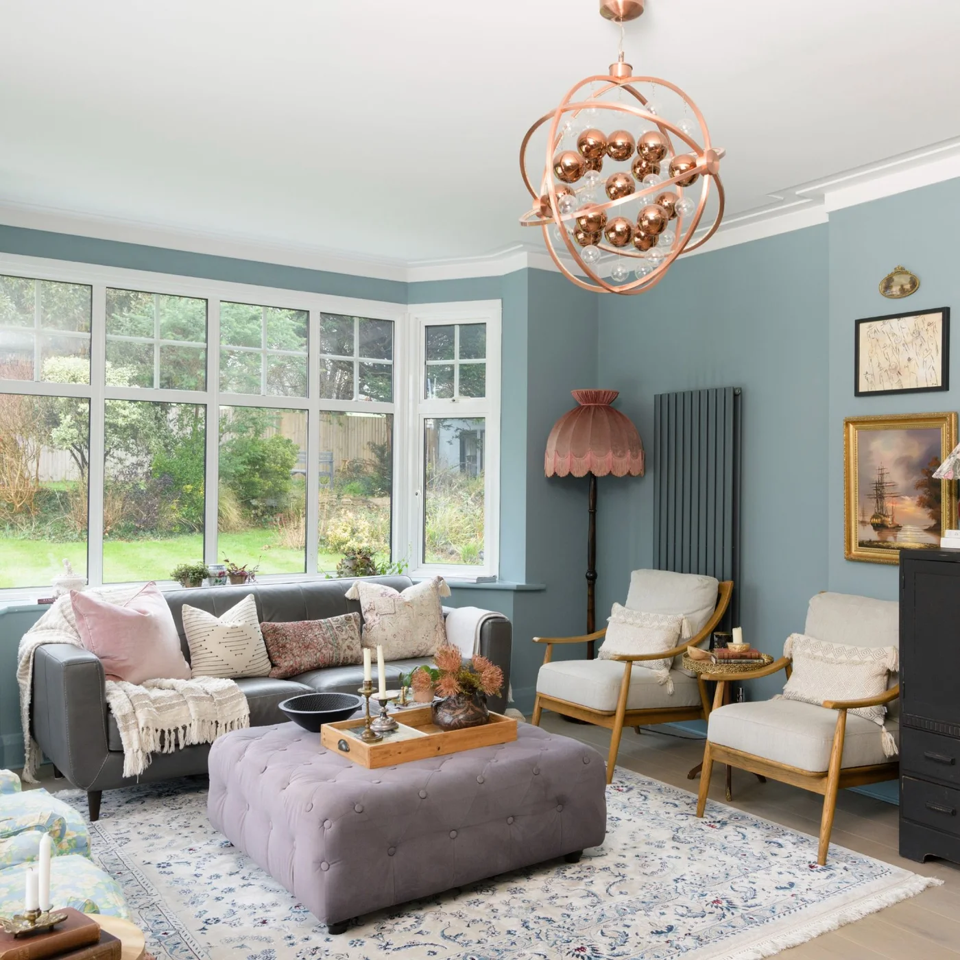 A dark gray sofa sits beneath broad windows opposite pale wooden-framed chairs around a tufted lilac-gray ottoman in a blue room