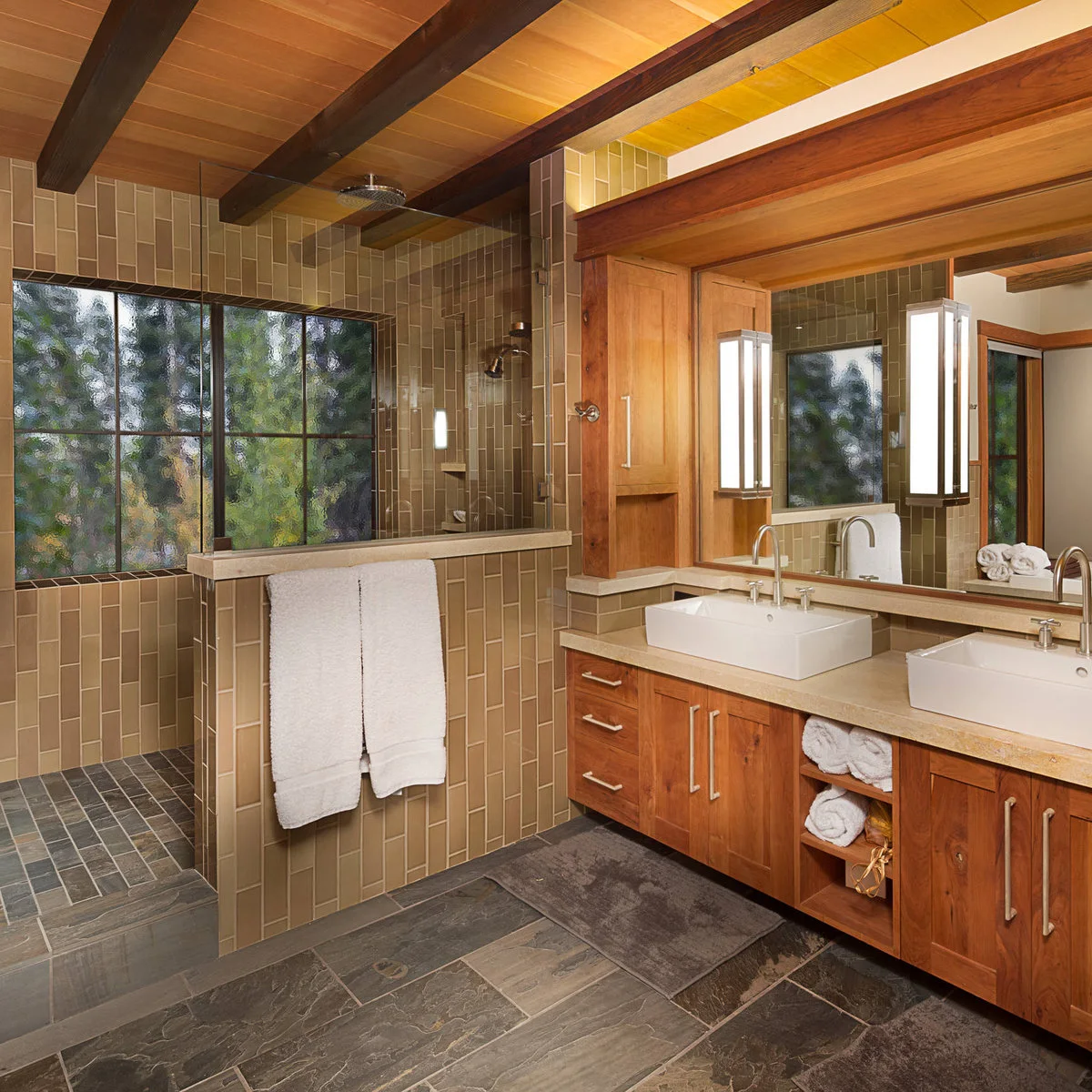 A shower with vertically arranged brown ceramic tiles, textured glazing and a half-height screen beside warm wood cabinetry.