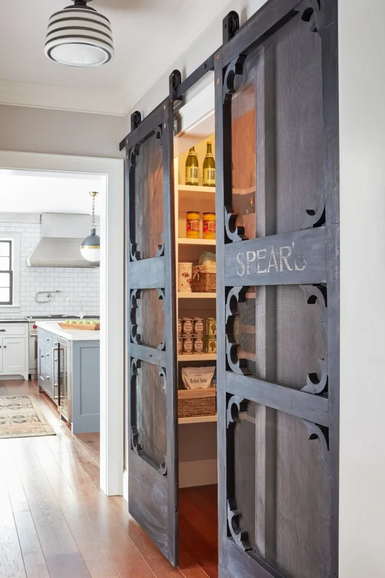 Dark sliding barn doors framing pantry shelves with jars and supplies beside pale kitchen cabinetry