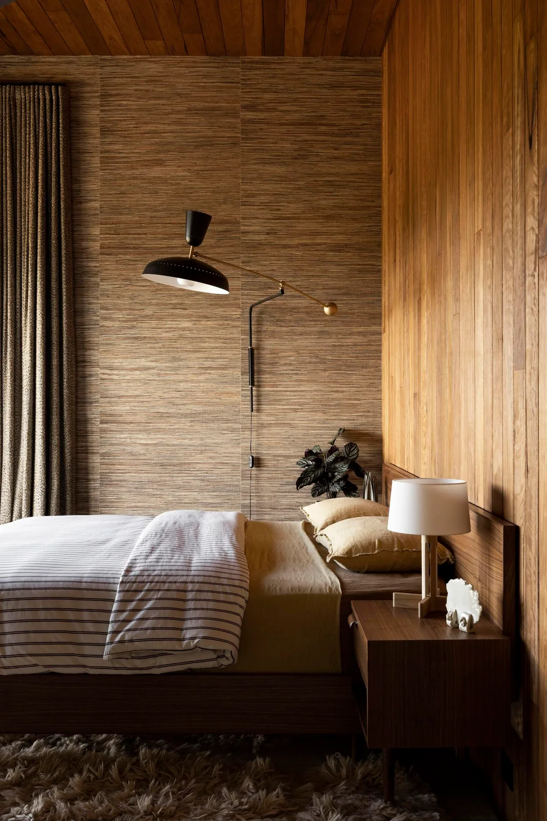 Fine striped bedding lies beneath textured bedroom walls and a wood-paneled ceiling beside a wood bedside cabinet, white table lamp and projecting dark wall light