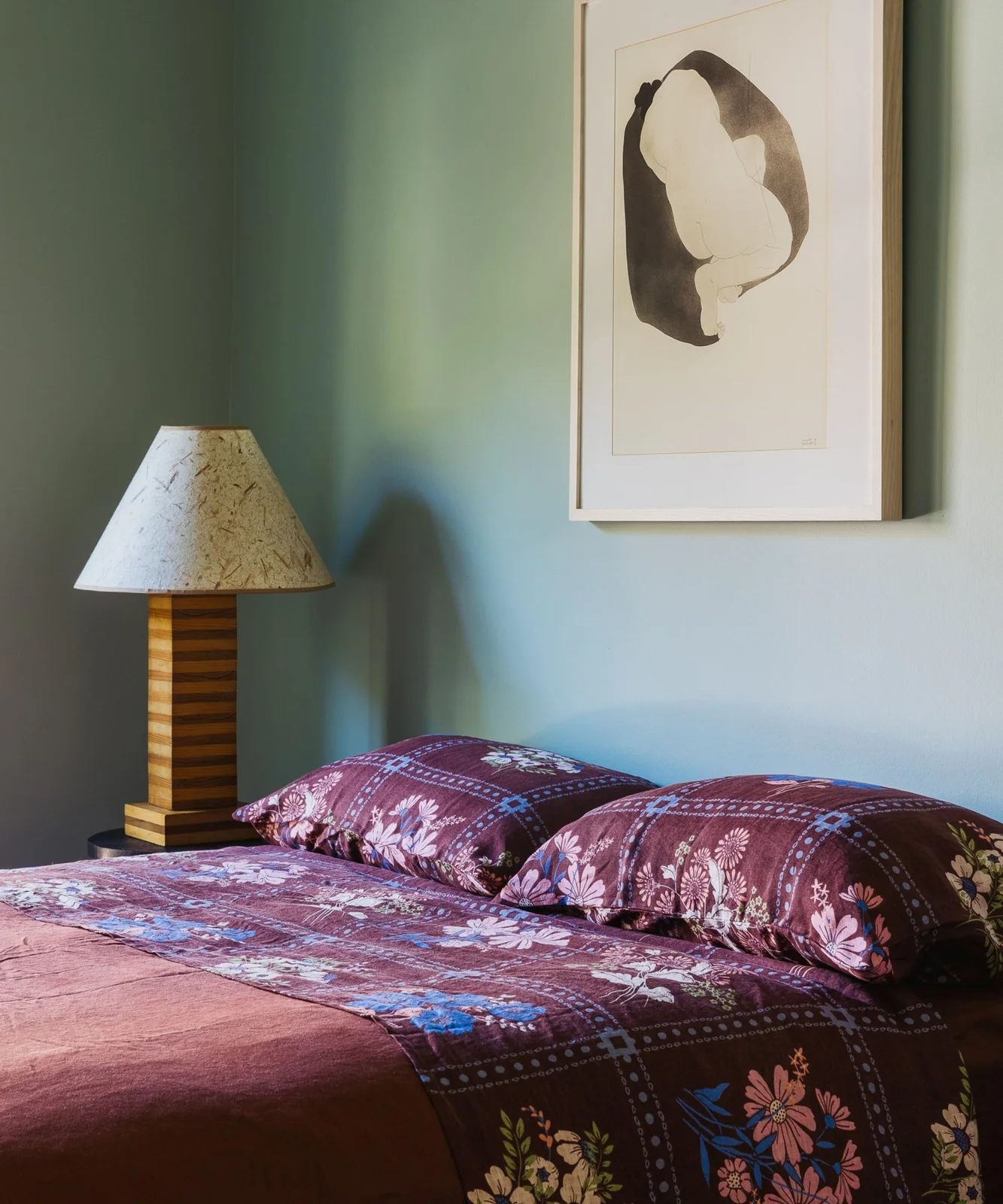 Pale green bedroom wall behind burgundy floral bedding with a wooden lamp and a light framed print.