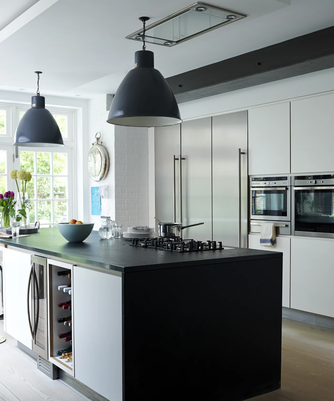 Flat pale cream kitchen cabinets with built-in ovens behind a dark counter island and large dark pendant lights.