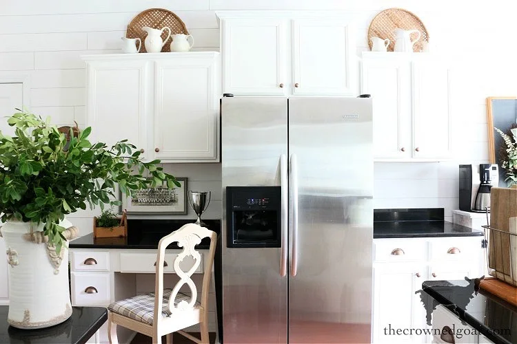 White pitchers arranged in front of round woven trays above white cabinets on both sides of a refrigerator.