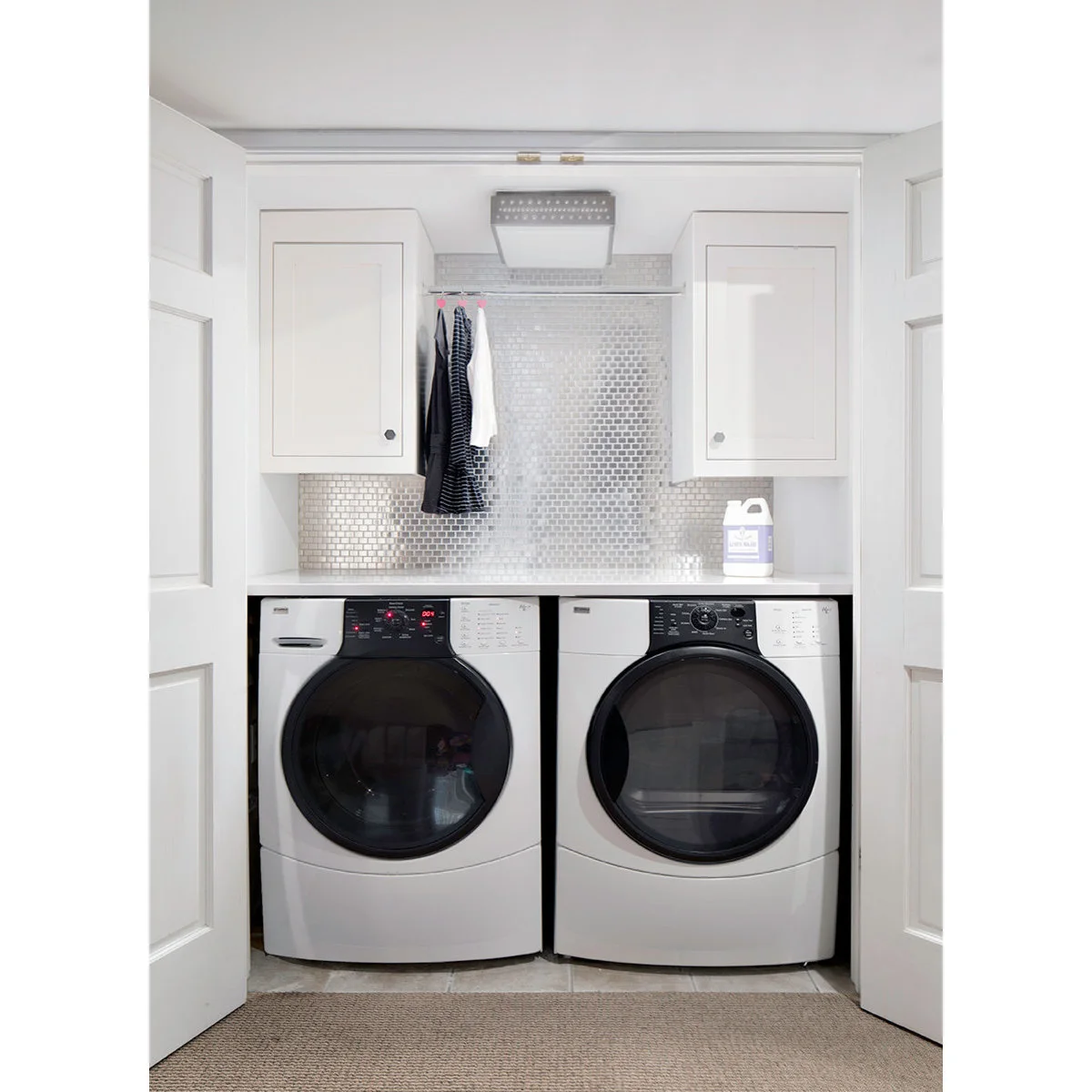 Open white laundry closet with side-by-side appliances, a counter, upper cabinets and a short central hanging rail