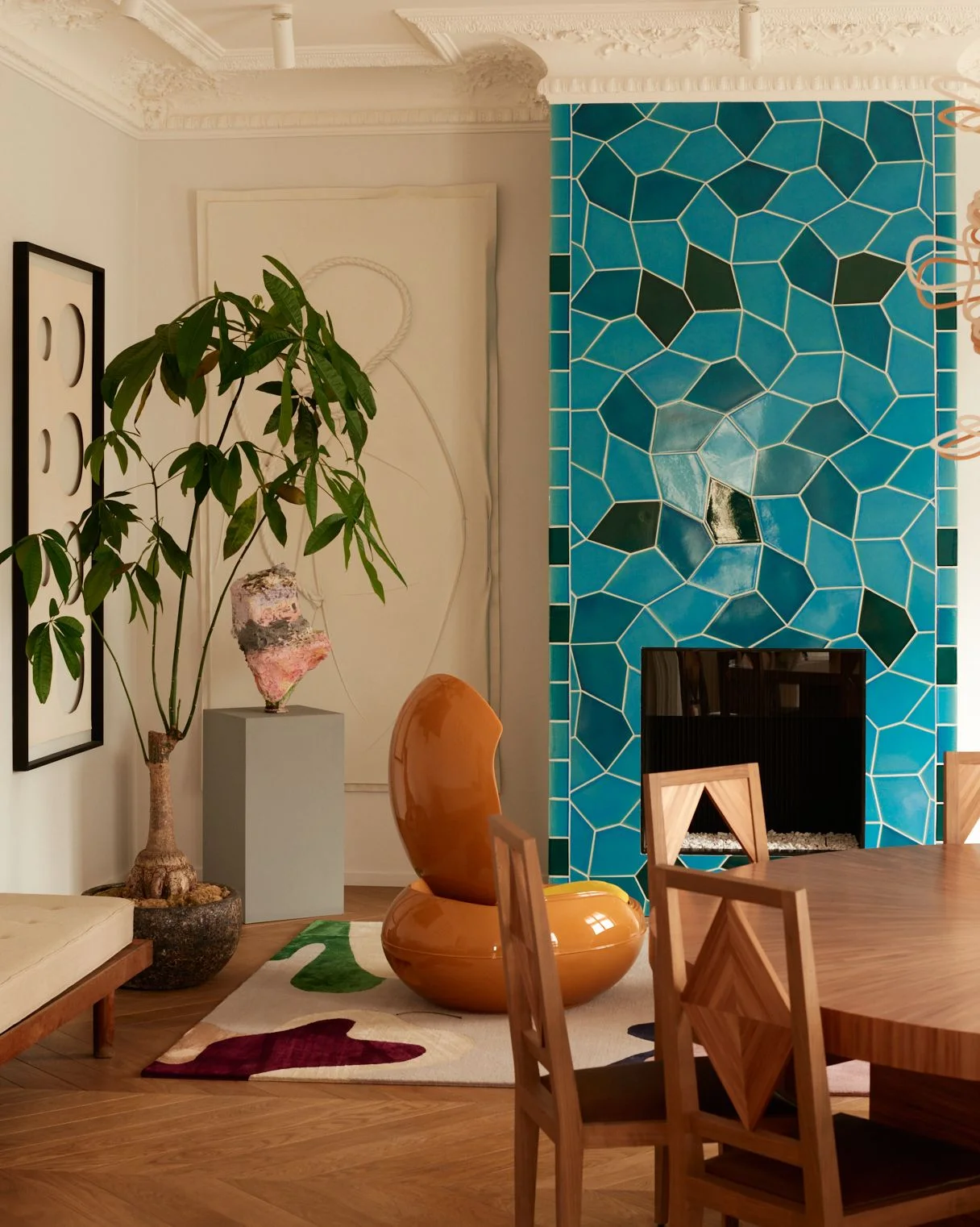 Irregular turquoise and dark green tiles with pale grout cover a tall fireplace beside an orange chair, a wood dining table and ornate pale molding.
