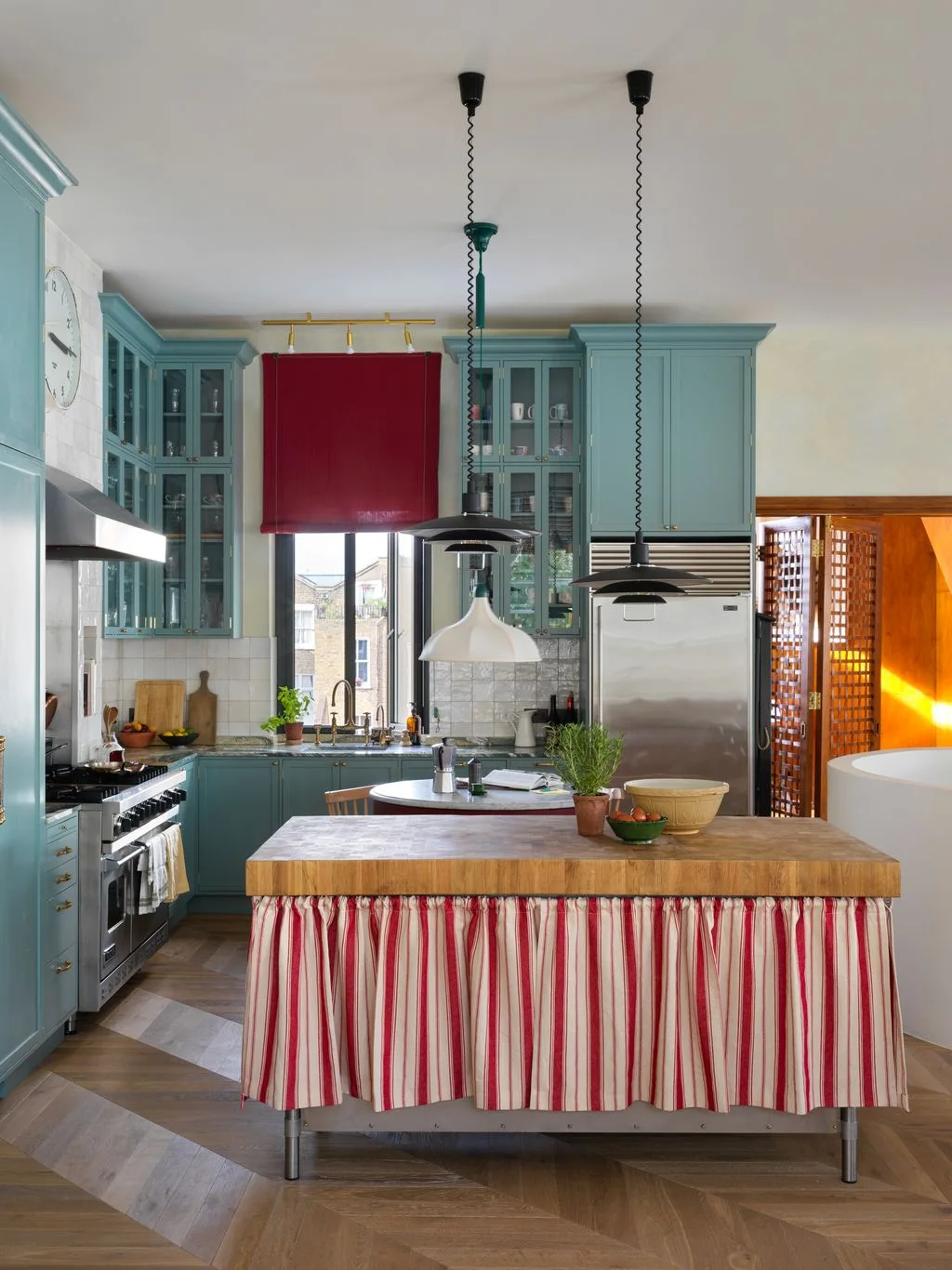 Blue kitchen cabinets surround a wood-topped island with a red-and-white striped skirt beneath black hanging lights beside a red window blind