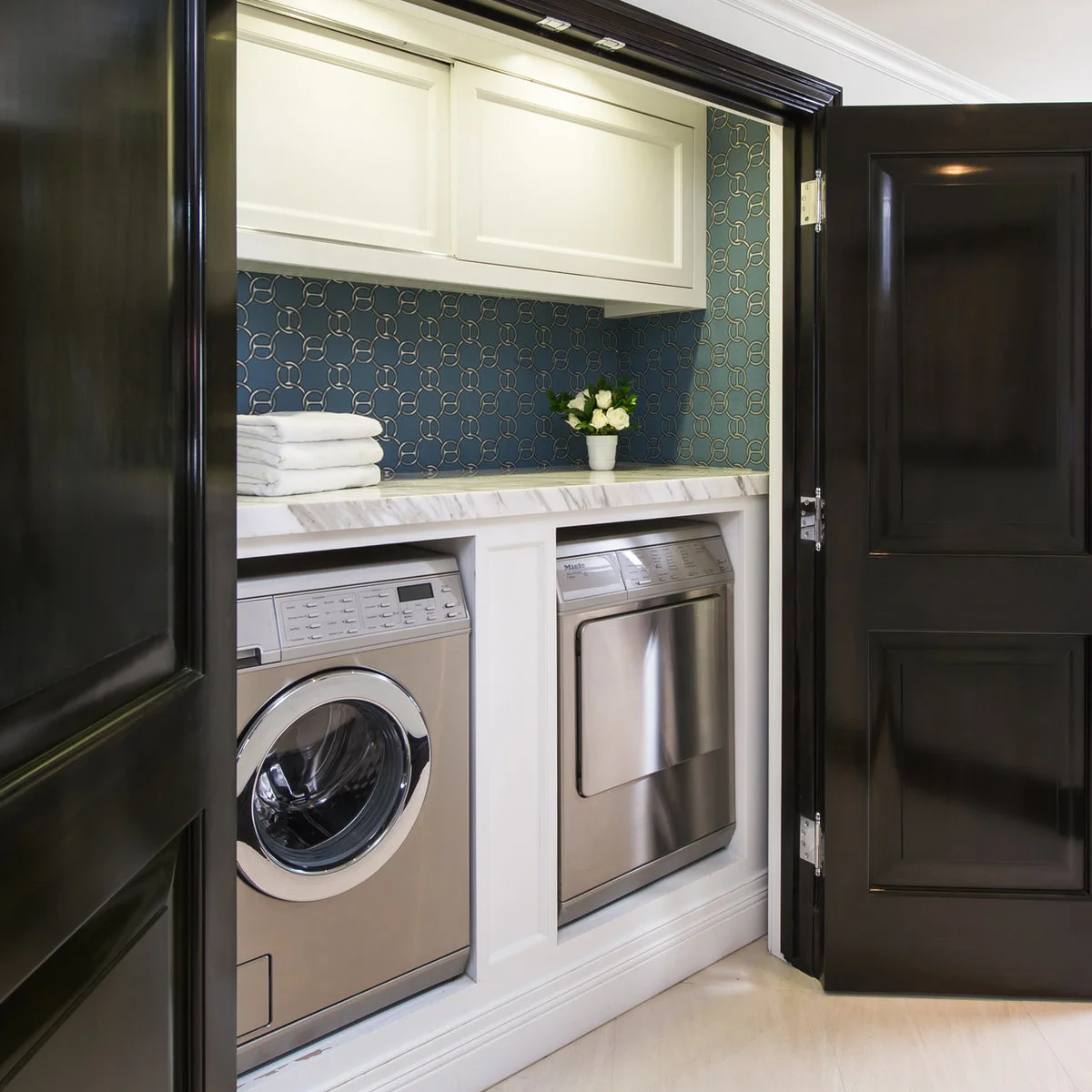 Laundry closet with black doors, a blue geometric backdrop, white cabinets and side-by-side appliances on a raised base