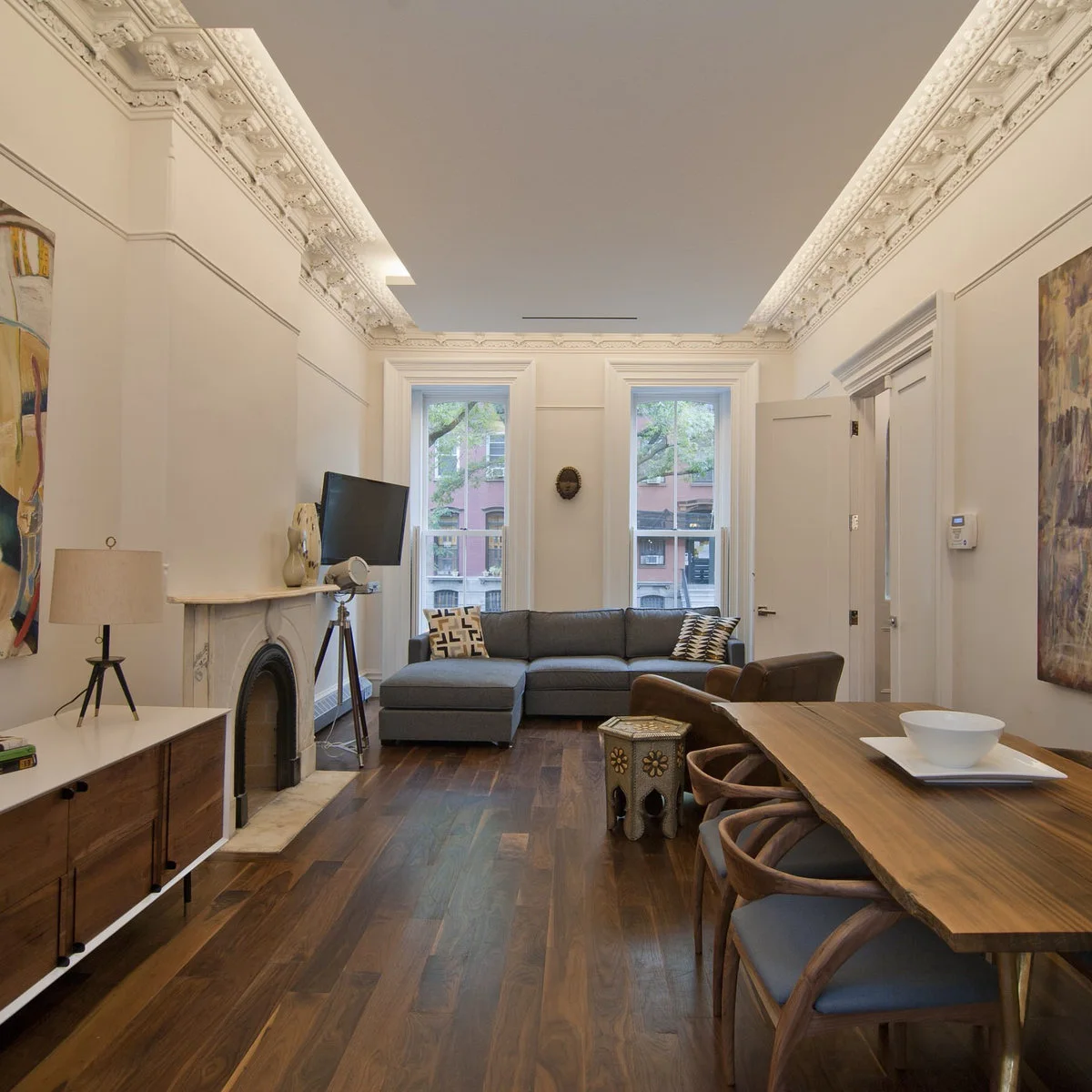 Long room with ornate pale ceiling trim, a gray sectional by two windows and a wooden dining table along the right wall.