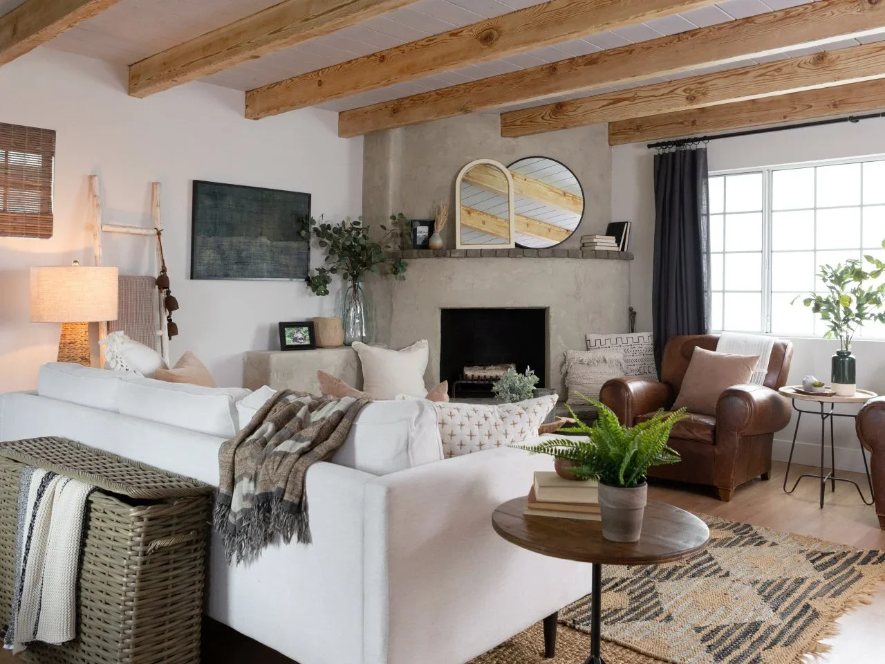 A white sofa with patterned cushions and a woven basket faces a pale fireplace beneath timber beams.