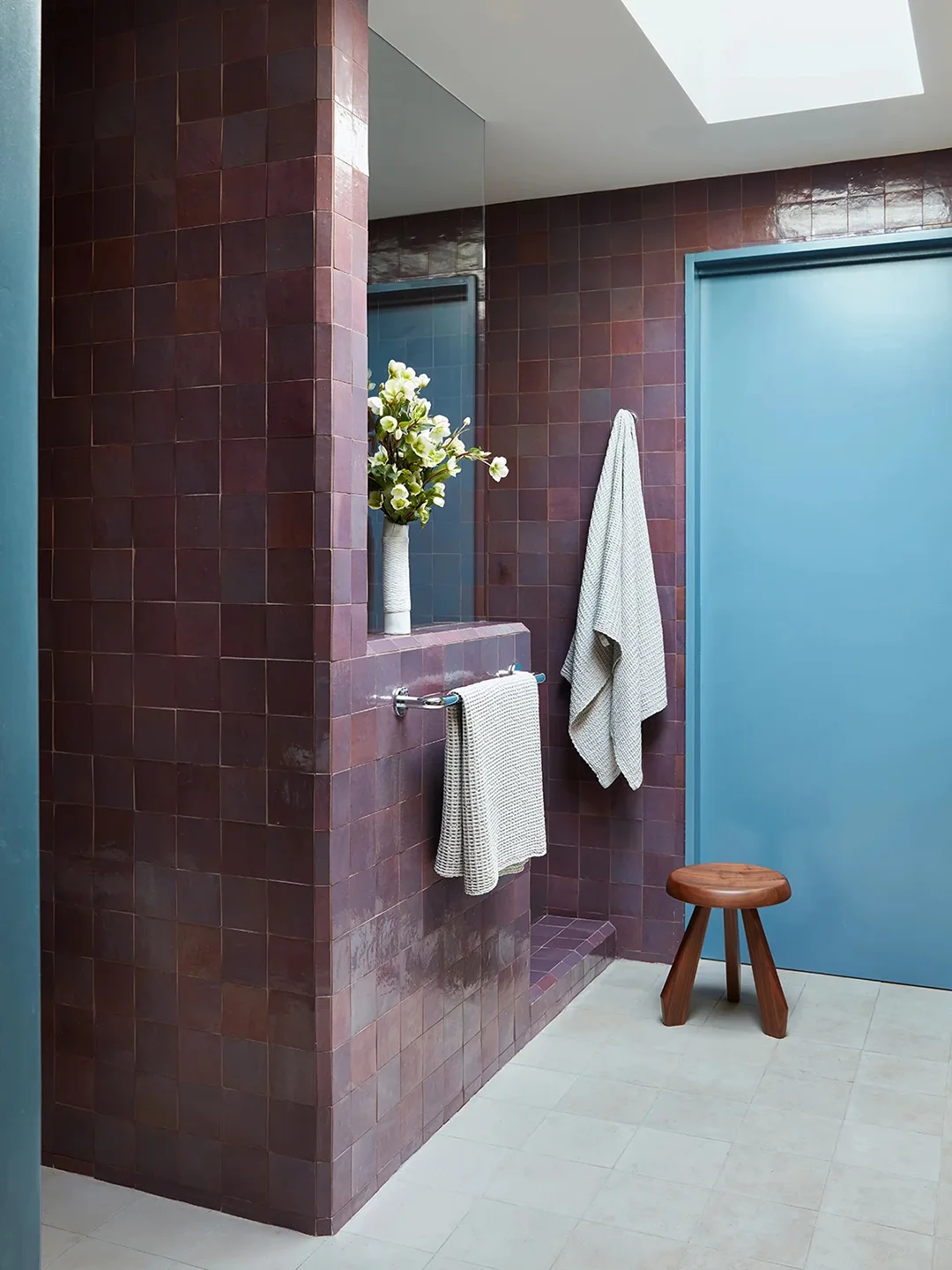 Glossy deep plum square bathroom tiles sit beside a blue door, a rounded wooden stool and a high glazed opening