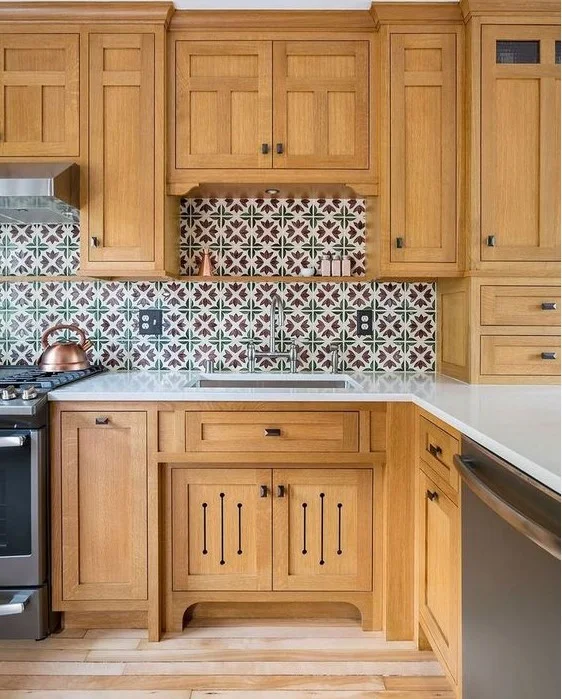 Warm wood-toned cabinets surround a pale counter and small sink beneath multicolored geometric patterned tiles with a tall storage front at the side