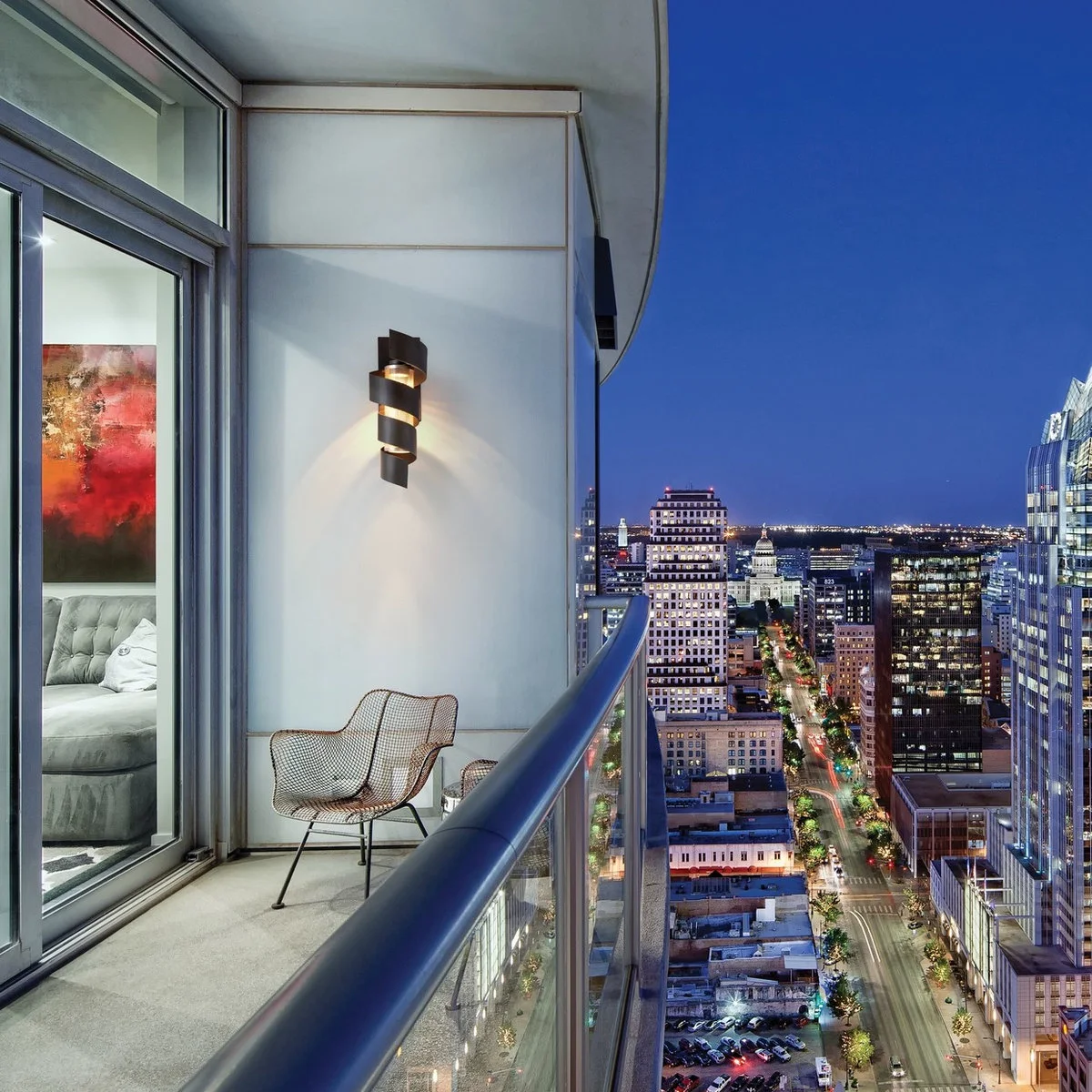 Narrow curved apartment balcony with one wire chair and a wall light at dusk