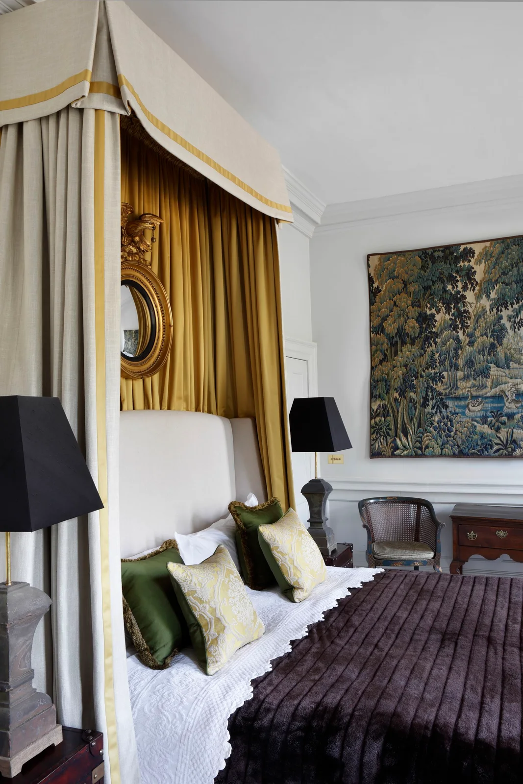 Mustard curtains rise behind a pale headboard beneath a cream fabric crown, with a plum bedcover, dark lamps and a gold-colored round mirror.
