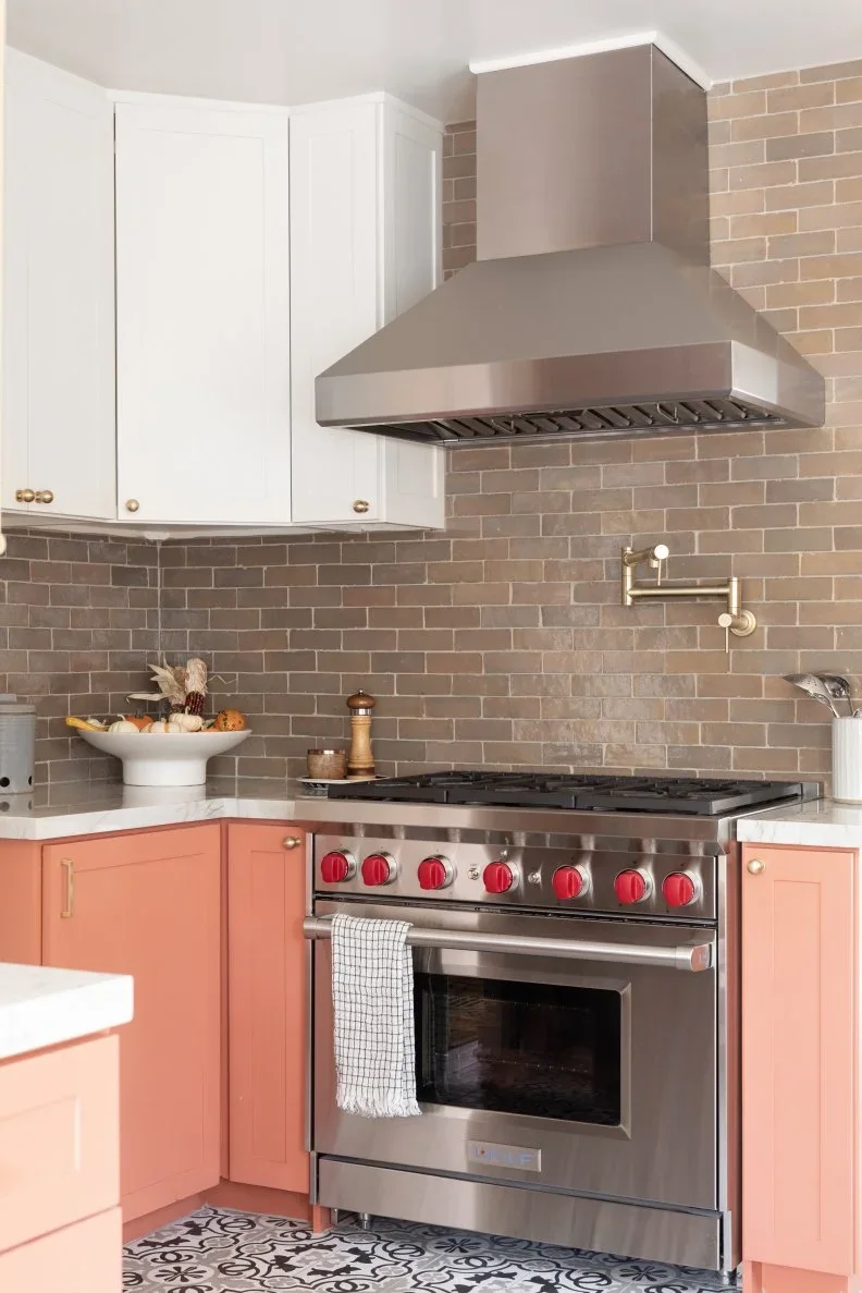 Coral pink lower cabinets and white uppers frame a stainless range beneath a metal hood and a warm neutral tiled backsplash.