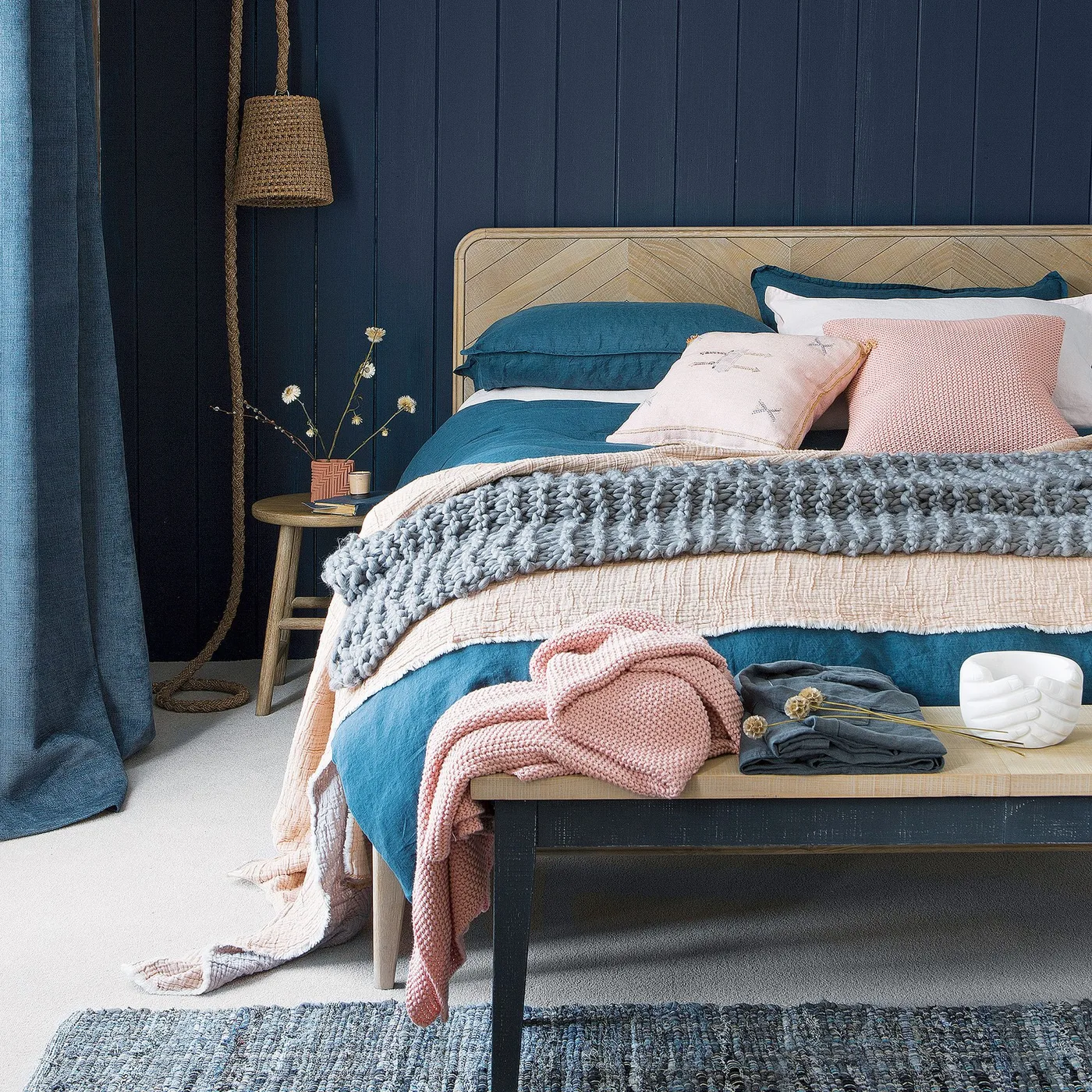 Navy paneled bedroom wall and curtains with a geometric wooden headboard, pink and gray throws, and a bench at the bed foot.