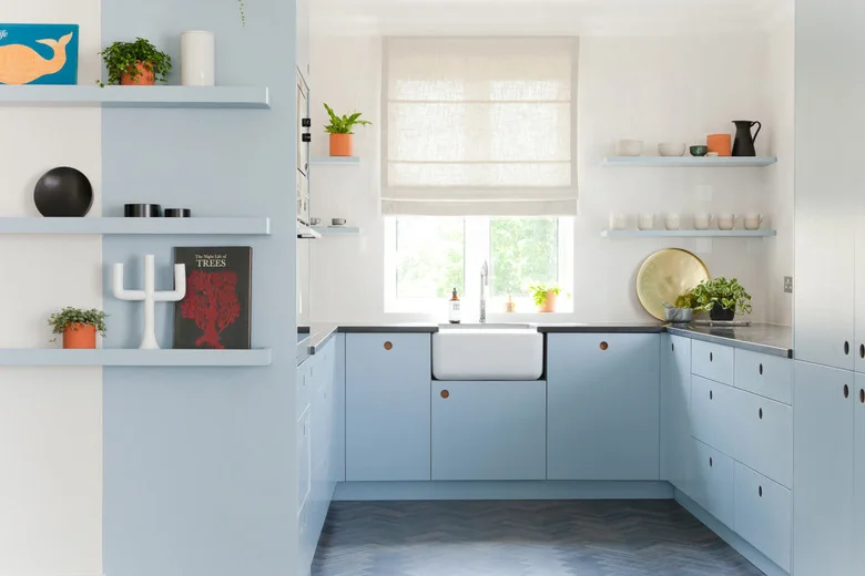 A pale blue U-shaped kitchen has flat doors with round wood pulls, an apron-front sink beneath a window, open shelves and a blue-gray patterned floor.