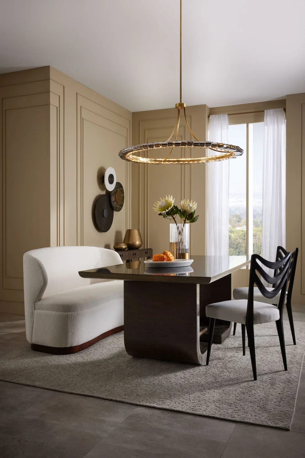 A curved pale upholstered dining bench sits beside a dark rectangular table and black chairs beneath a ring pendant against muted paneled walls and pale curtains