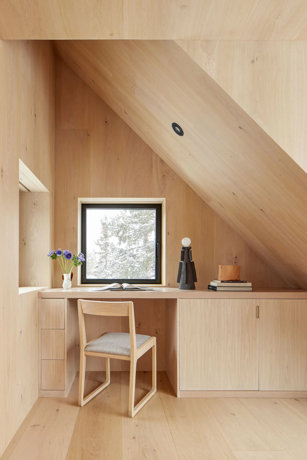 A pale wood-lined attic nook has an integrated desk beneath a small window, drawers at one side, sliding storage at the other and a simple wood chair.