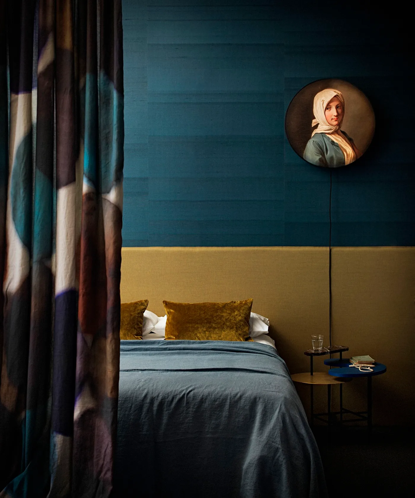Deep teal bedroom wall with an ochre headboard and cushion, dark patterned curtain, round portrait, and compact bedside table.