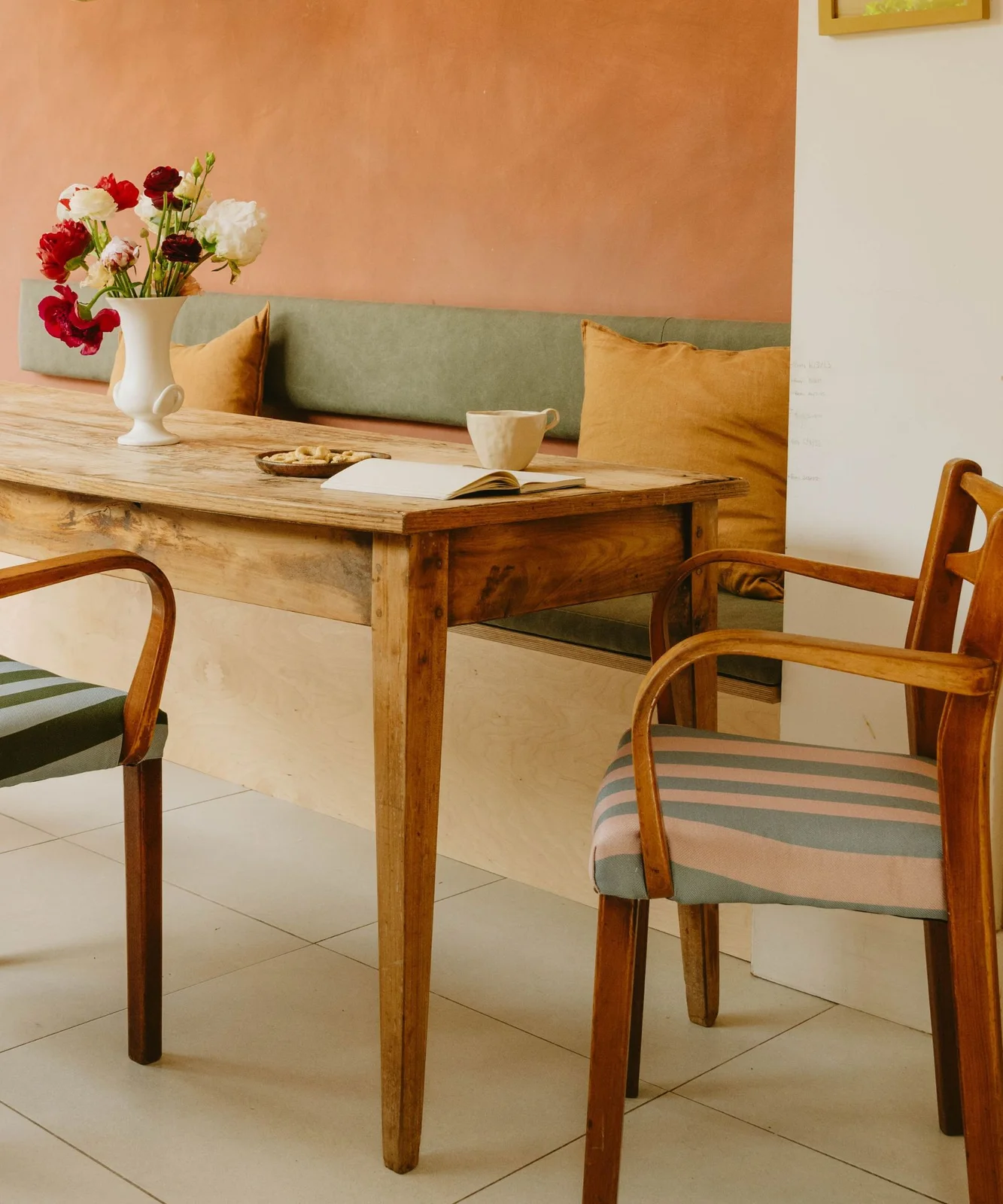 Wood dining chairs with pale green and pink striped seats sit beside a simple wood table and green banquette beneath warm orange walls