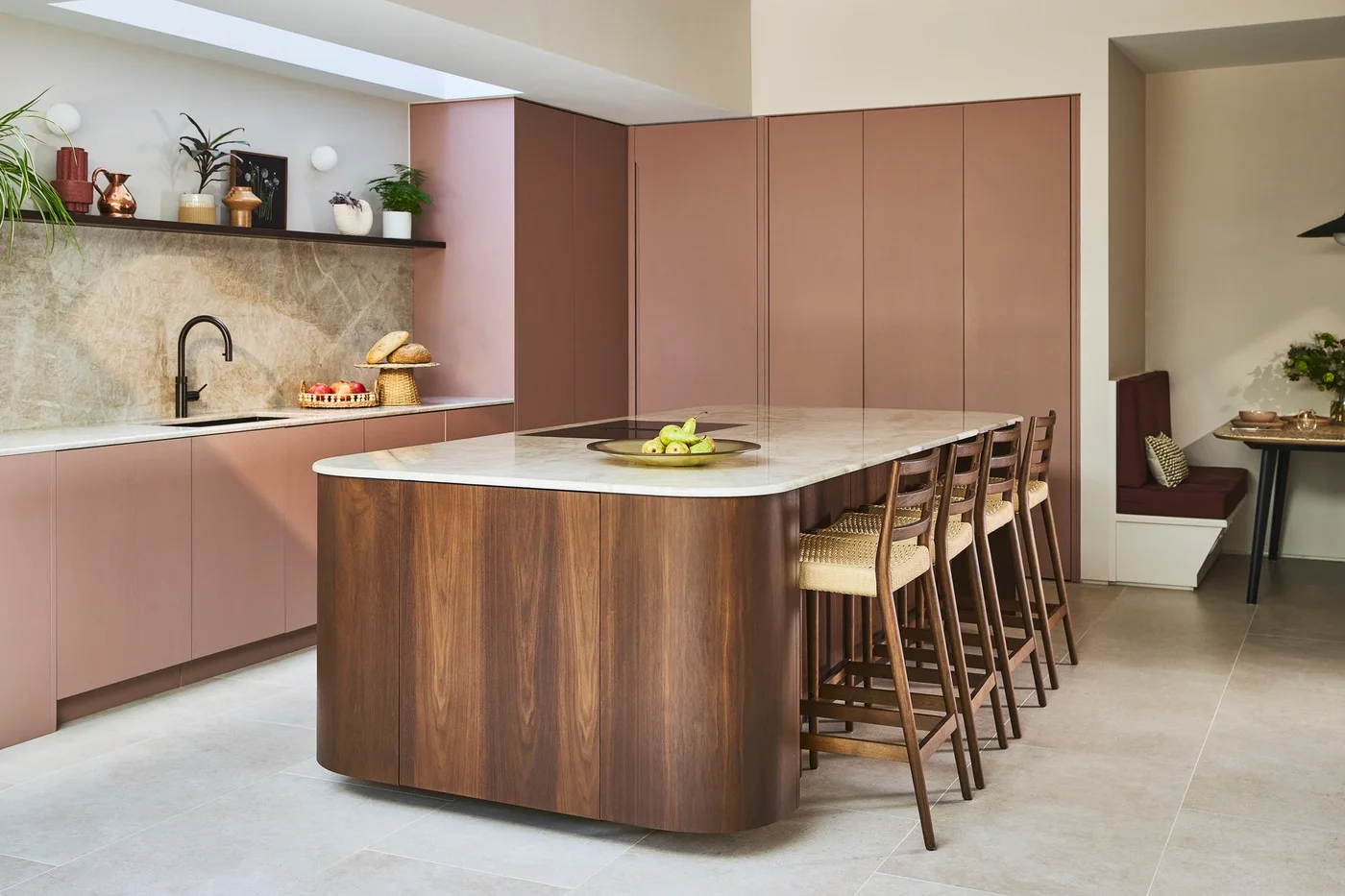 A curved walnut island with woven stools stands beside dusty pink cabinets and a pale veined backsplash.