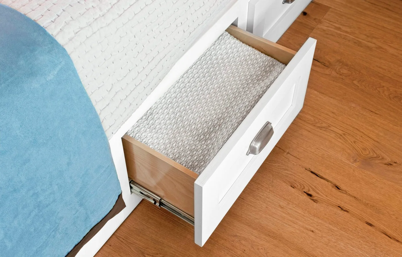 A white drawer pulls out from beneath a guest bed, holding neatly folded pale bedding beside a wood floor.
