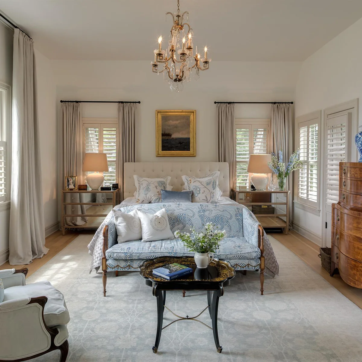Pale bedroom with patterned settee at the bed foot, chandelier and curved wood-colored chest